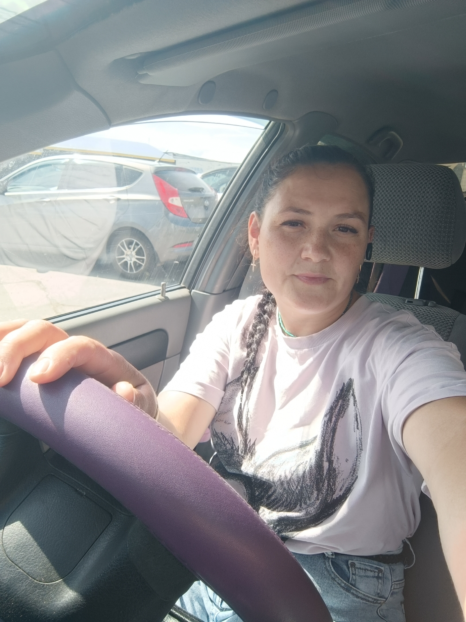 A selfie taken from inside a car in Chaikovsky, Russia. The image features a woman with dark hair pulled back in a braid, wearing a light-colored t-shirt with a graphic print and blue jeans. She is holding the steering wheel, which is covered in a purple material. Another car is visible through the car window, partially covered by a tarp. The scene appears to be outdoors, with sunlight illuminating the interior of the car.