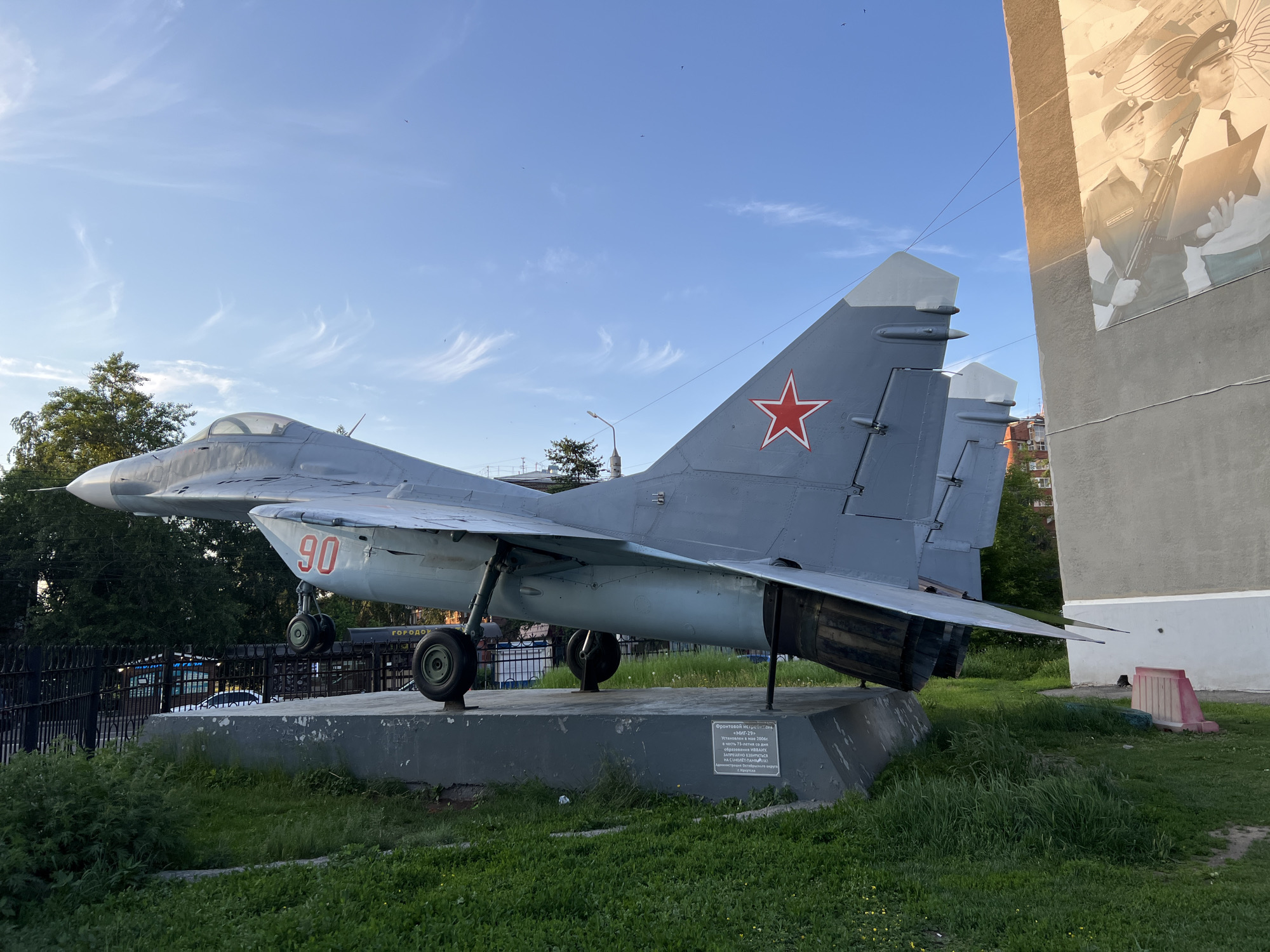 The image depicts a Mikoyan MiG-29 fighter jet displayed as a monument on a concrete pedestal in Irkutsk, Russia. The jet is painted gray and features a red star emblem on its tail fin and the number "90" in red on its fuselage. It is positioned outdoors on a grassy area, with trees and a building in the background. The building has a mural on its side depicting two figures in military attire. The sky is clear with some wispy clouds. There is a plaque on the pedestal with Russian text, which appears to be a dedication or information about the aircraft. The overall scene is static, with no people or animals visible. The lighting suggests it is daytime, likely late afternoon due to the angle of the sun.