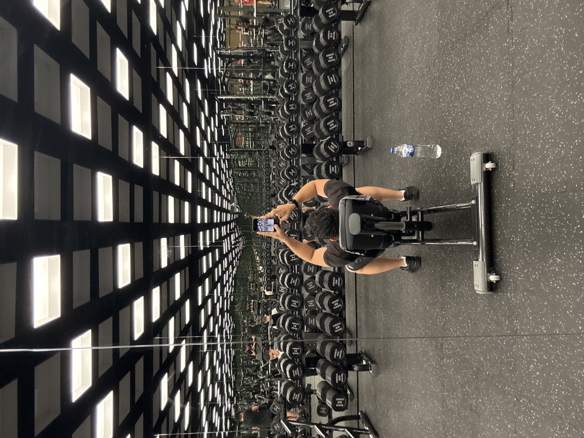 A person is taking a selfie in a well-lit gym with mirrors on all sides. The person is seated on a weight bench, facing away from the camera, and holding a smartphone in their right hand to capture the reflection. The gym is equipped with numerous dumbbell racks filled with weights of various sizes. The ceiling features a grid of square fluorescent lights. In the foreground, a water bottle rests on the rubberized floor. The scene is a gym in Varna, Bulgaria.