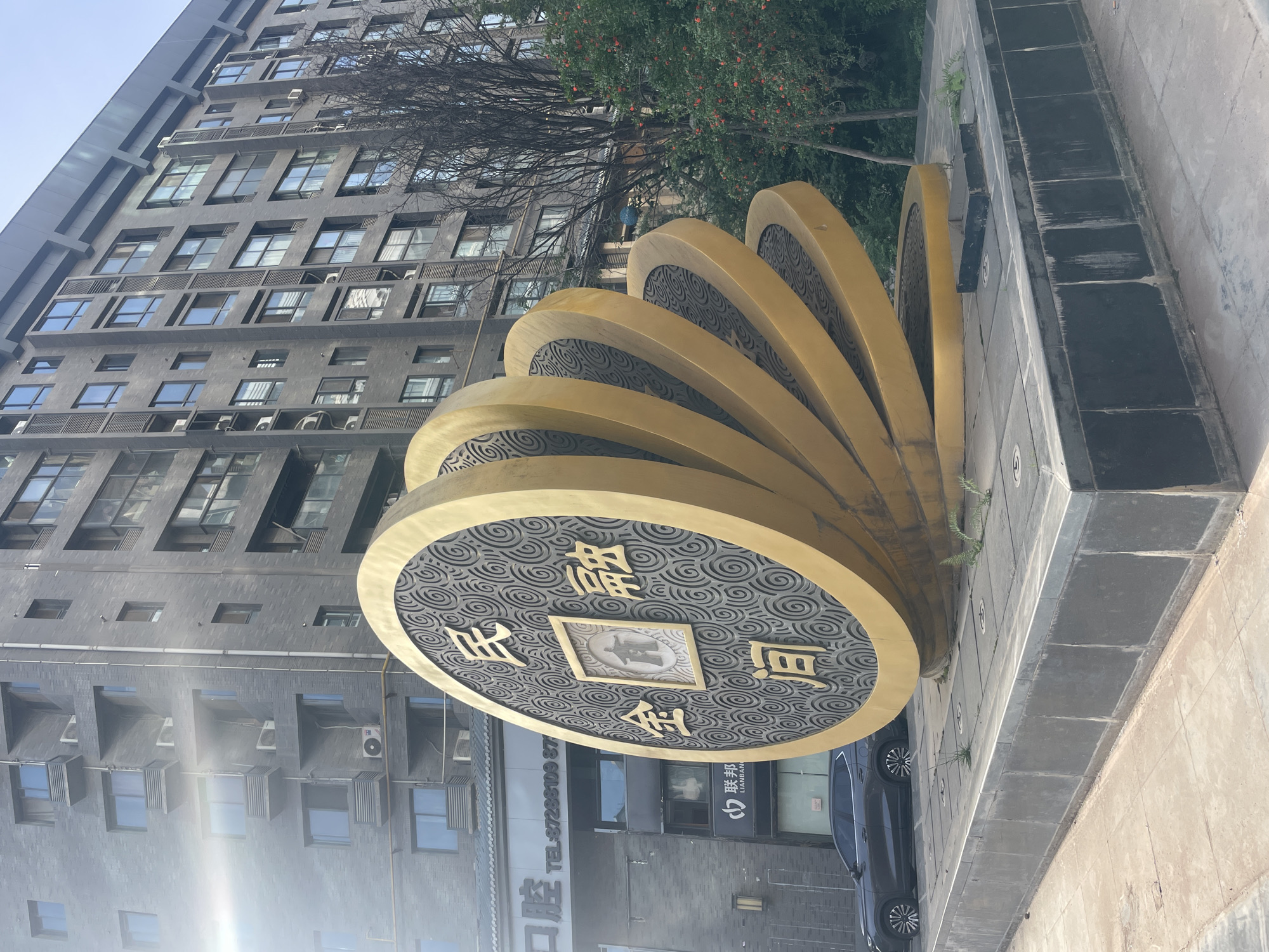 The image shows a large, golden, coin-shaped sculpture in Xincheng District, China. The sculpture is situated on a raised platform with dark gray paving stones. The coin faces left and has a decorative pattern on its surface with four Chinese characters and a rectangular frame at its center. A dark gray building rises in the background, and there is a vehicle parked near the sculpture.
