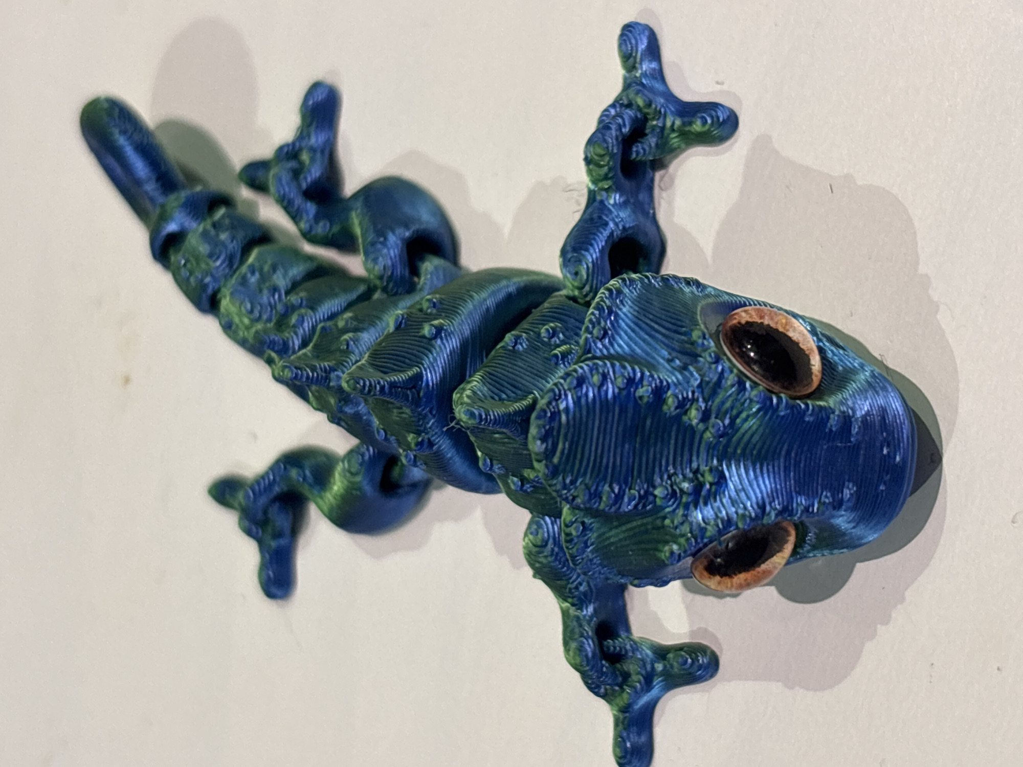An overhead shot shows a blue and green 3D printed articulated gecko toy with orange eyes on a white surface. The toy has a segmented body and legs that allow for articulation, and its surface texture reveals the layered printing process. The gecko is positioned centrally on the surface, with its head facing towards the viewer and its tail extending upwards and to the right. Shadows cast by the toy indicate a light source from the upper left. No specific location in Dubai, United Arab Emirates, is identifiable from the image.