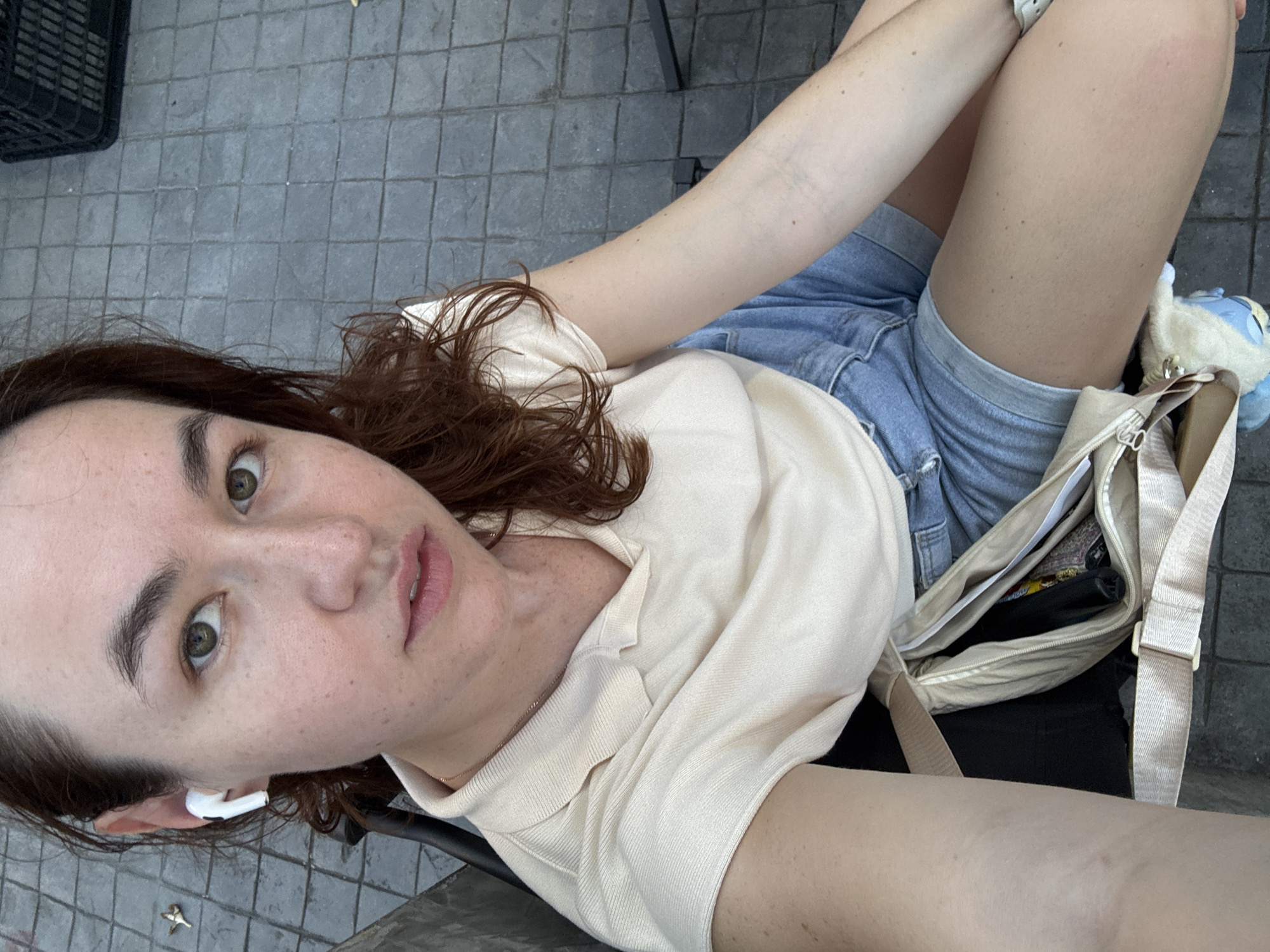 A young woman with brown hair and green eyes is seated outdoors on a tiled surface. She is wearing a light-colored polo shirt and denim shorts. A light-colored crossbody bag is slung across her body. She has a white earbud in her left ear. The background shows a grey tiled ground and part of a black plastic crate. The scene does not contain any specific elements indicating it is located in Surat Thani City Municipality, Thailand.