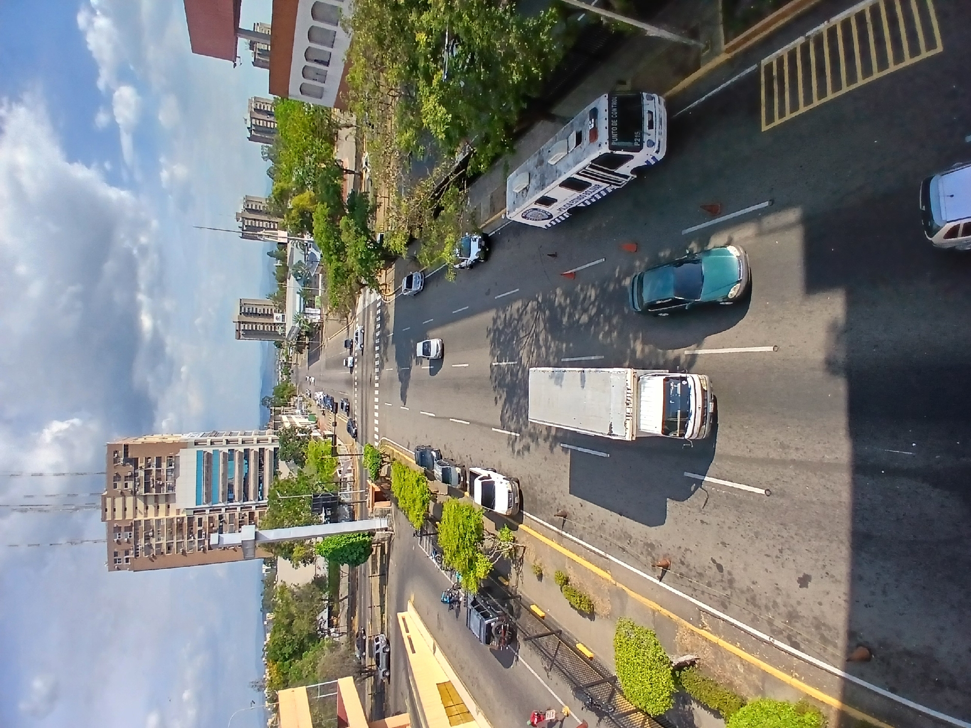 The image depicts a view from above a multi-lane road in Ciudad Guayana, Venezuela. Several cars, a large white truck, and a bus marked "PUNTO DE CONTROL POLICIA ESTADO BOLIVAR" are visible on the road. The bus is positioned near the edge of the road. Buildings, trees, and sidewalks line the sides of the road. The sky is partly cloudy.