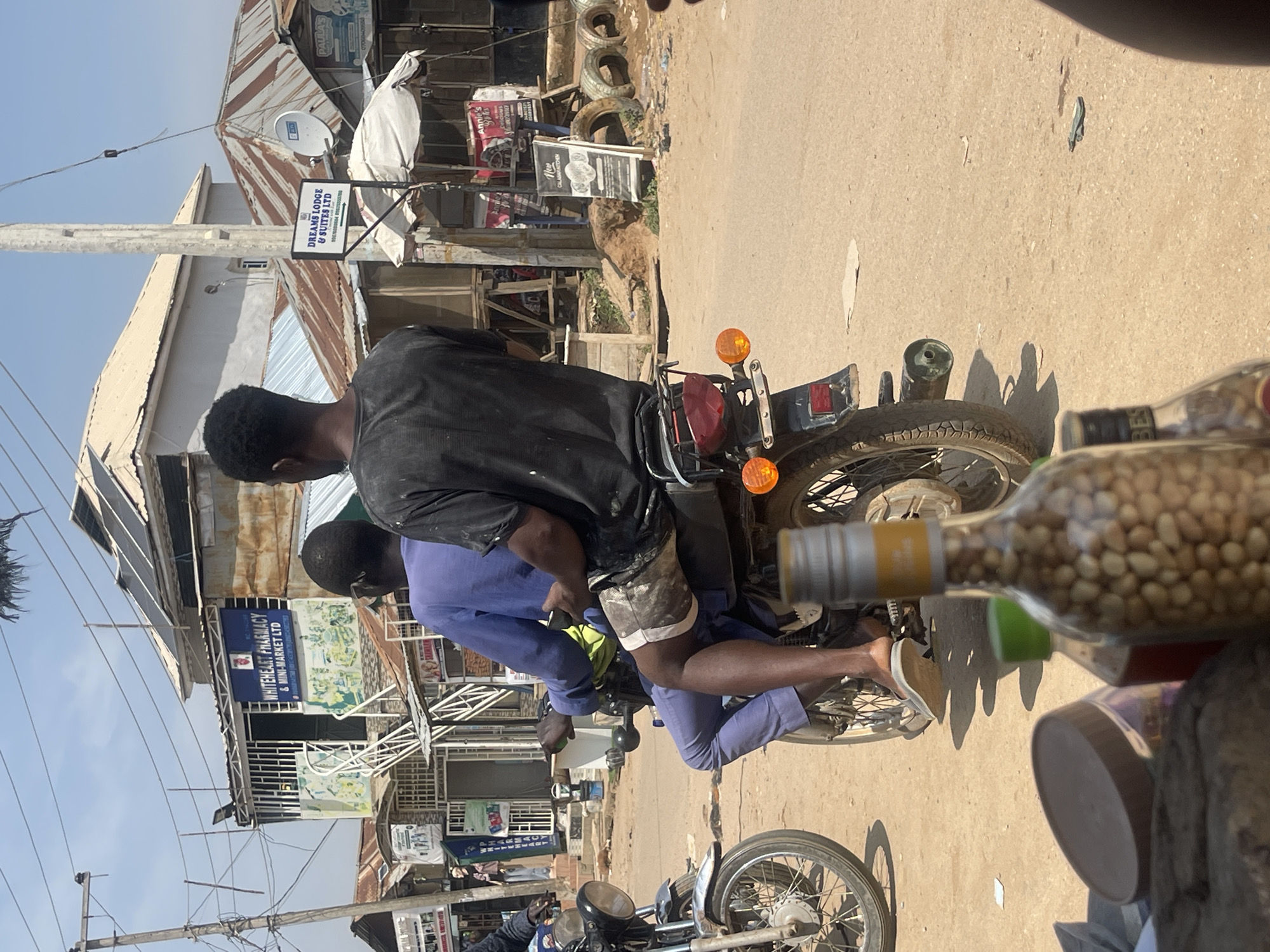 In an outdoor urban scene in Abuja, Nigeria, two men ride a motorcycle on a dirt road. The man in front, wearing a blue outfit, is driving, while the man behind him, wearing a black t-shirt, is a passenger. The motorcycle has a license plate. In the background, there are several buildings with corrugated metal roofs, some of which appear to be shops or businesses. One sign reads "WHITEHEART PHARMACY & MINI-MARKET LTD", and another reads "DREAMS LODGE & SUITES LTD". There are also power lines and utility poles visible. In the foreground, there are several bottles, one of which is filled with peanuts. The scene appears to be a typical street view in a developing urban area.