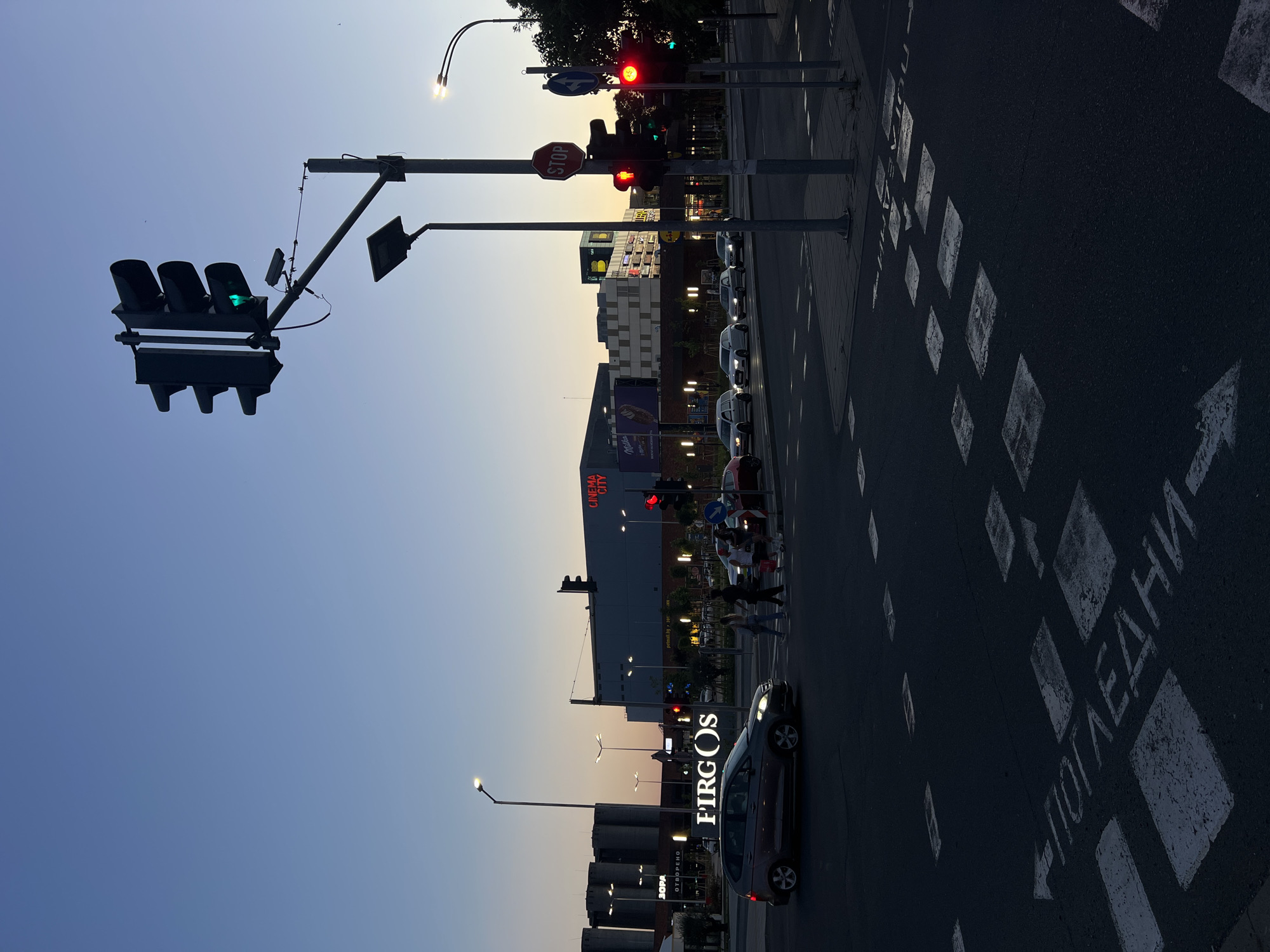 The image depicts a street intersection in Burgas, Bulgaria, during twilight. A four-way traffic light is illuminated with a green light for vehicles. Across the street, a large building with "CINEMA CITY" signage is visible. Several cars are present, with one dark-colored hatchback in the foreground and another red car to the right. A group of pedestrians is crossing the street on a marked crosswalk. The road surface features white painted crosswalk markings and directional arrows indicating traffic flow. The sky is a clear, pale blue, transitioning to a darker hue towards the horizon, indicating either sunrise or sunset. Streetlights are also illuminated.