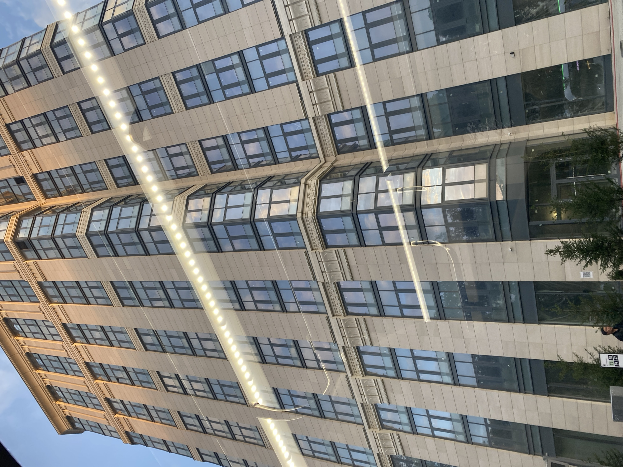 The image captures a modern, tall building with a light-colored facade and numerous windows. A string of lights runs diagonally across the frame, creating a reflection on the building's surface. Several trees are visible at the base of the building. In the lower left corner, a man stands near a bus stop sign. The scene appears to be an urban environment. Based on the visual cues like the architectural style and the presence of a bus stop, this location is likely in Moscow, Russia.
