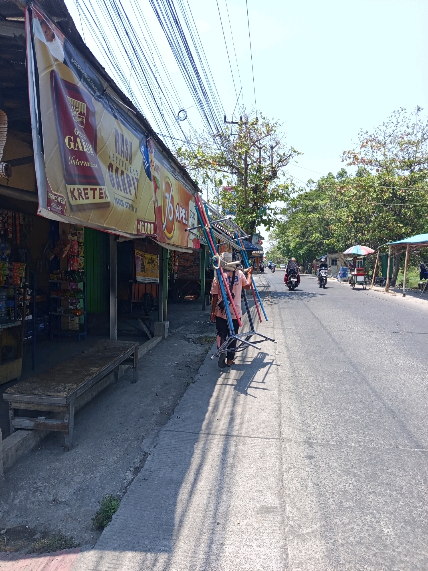 The image depicts a street scene in Karawang, Indonesia. A person wearing a pink shirt and a white hat is walking along the side of a road, carrying a disassembled metal display rack. The rack appears to be made of blue, red, and yellow metal poles. To the left of the person is a small shop with a banner advertising "GAJAH" cigarettes. Shelves stocked with various items are visible inside the shop, and a wooden bench sits outside. The road is paved, with vehicles, including motorcycles, visible in the distance. Trees line the side of the road, and utility poles with numerous wires are prominent in the background. The sky is bright and clear.