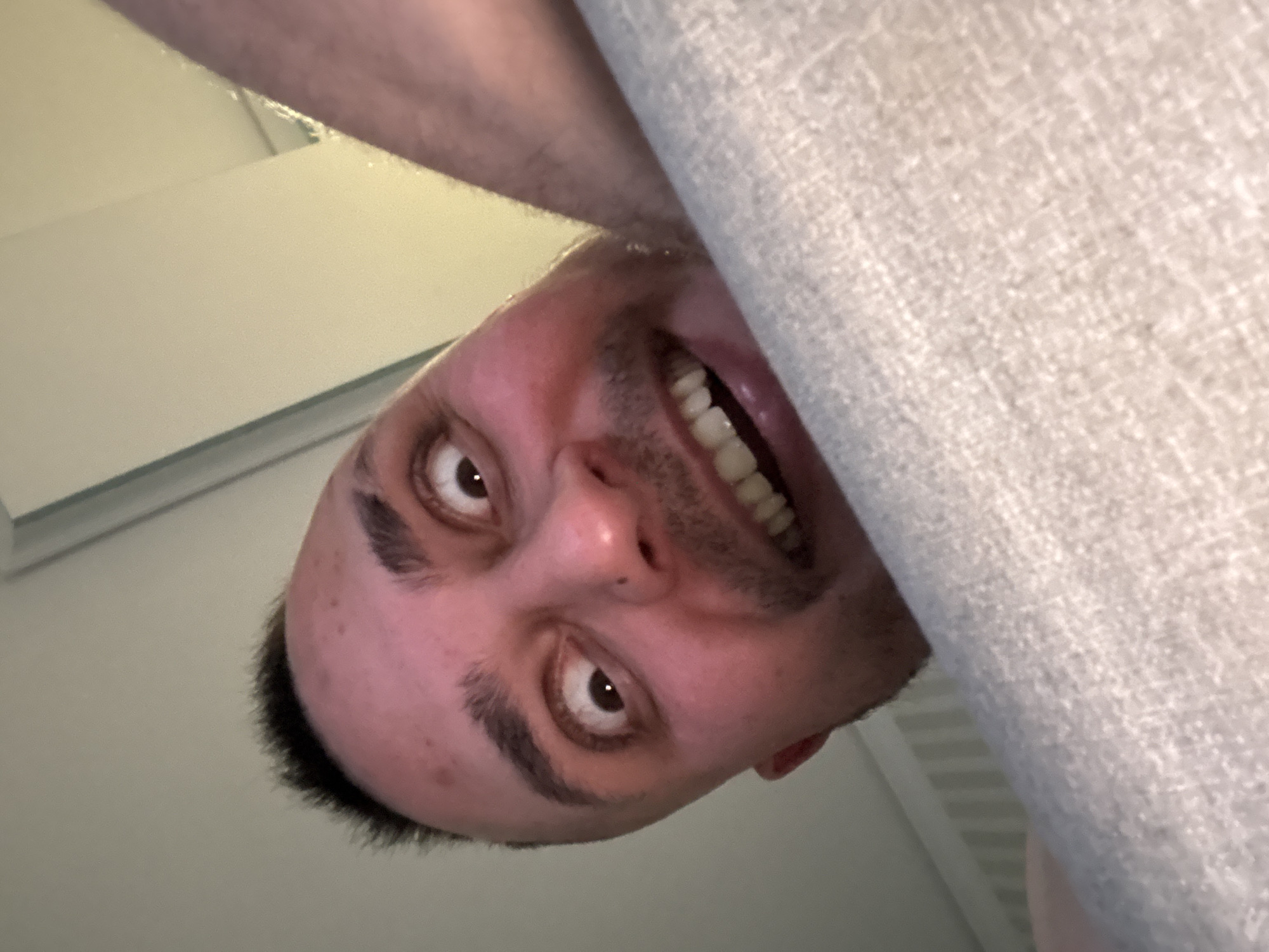 A close-up, upside-down selfie of a Caucasian male with a shaved head, dark hair, and a light complexion. He is smiling, revealing his teeth. Part of a light grey, textured fabric or object is prominently positioned to the right of his face. In the upper left corner, a white or light-colored, possibly manufactured surface is visible. The location and city are not discernible.