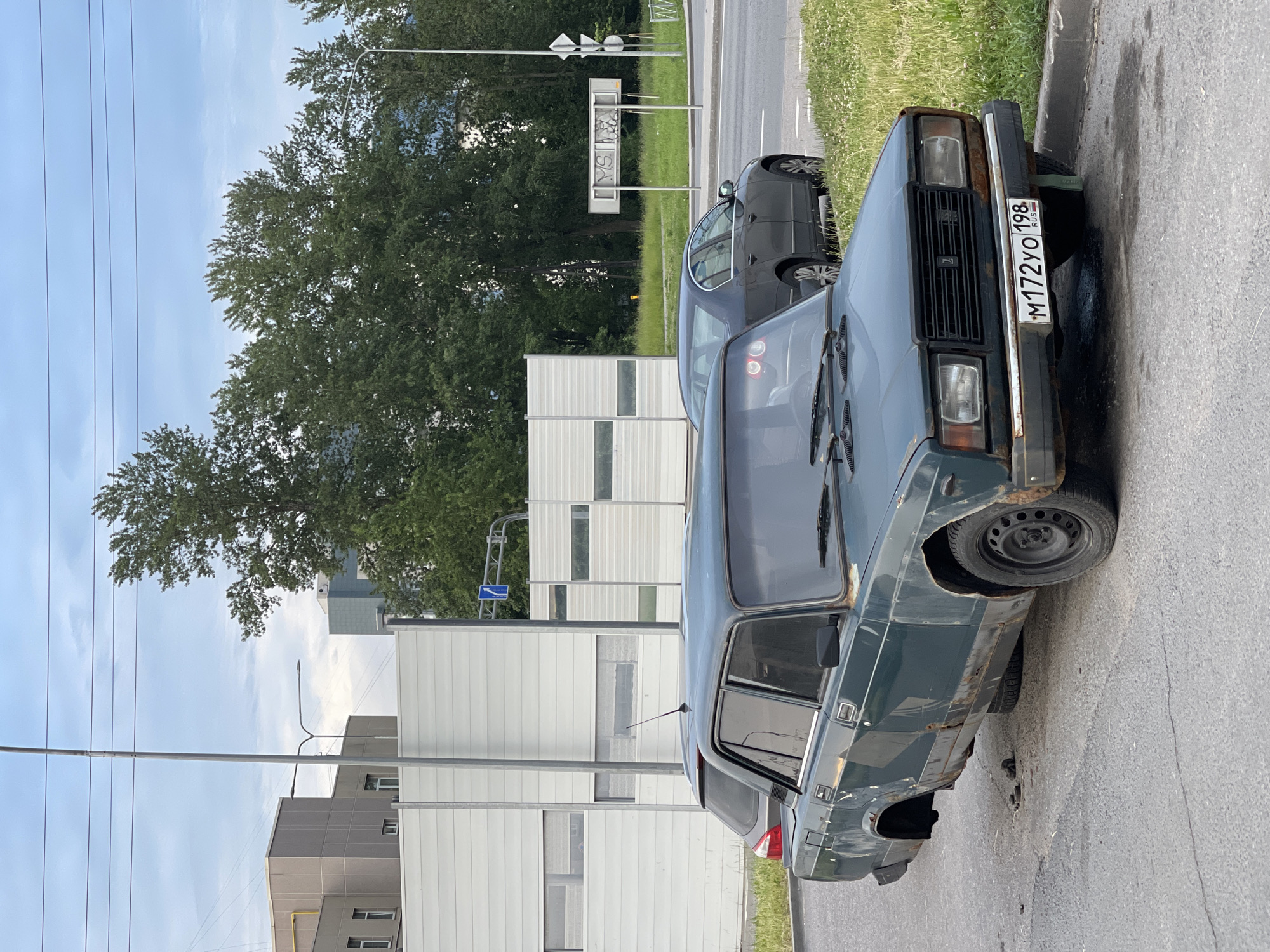 A dark green, heavily rusted sedan, identified by its license plate "M172YO 198 RUS," is parked on a paved surface, with its rear passenger-side wheel and suspension damaged, causing the vehicle to tilt. A spare tire is visible underneath the car. The car appears to be a Lada, given its design. In the background, a darker colored sedan is parked on a grassy area next to a road. Beyond the road, a modern white sound barrier separates the street from a grassy area with trees and several buildings. The sky is overcast. The scene is in a seemingly urban or suburban environment.