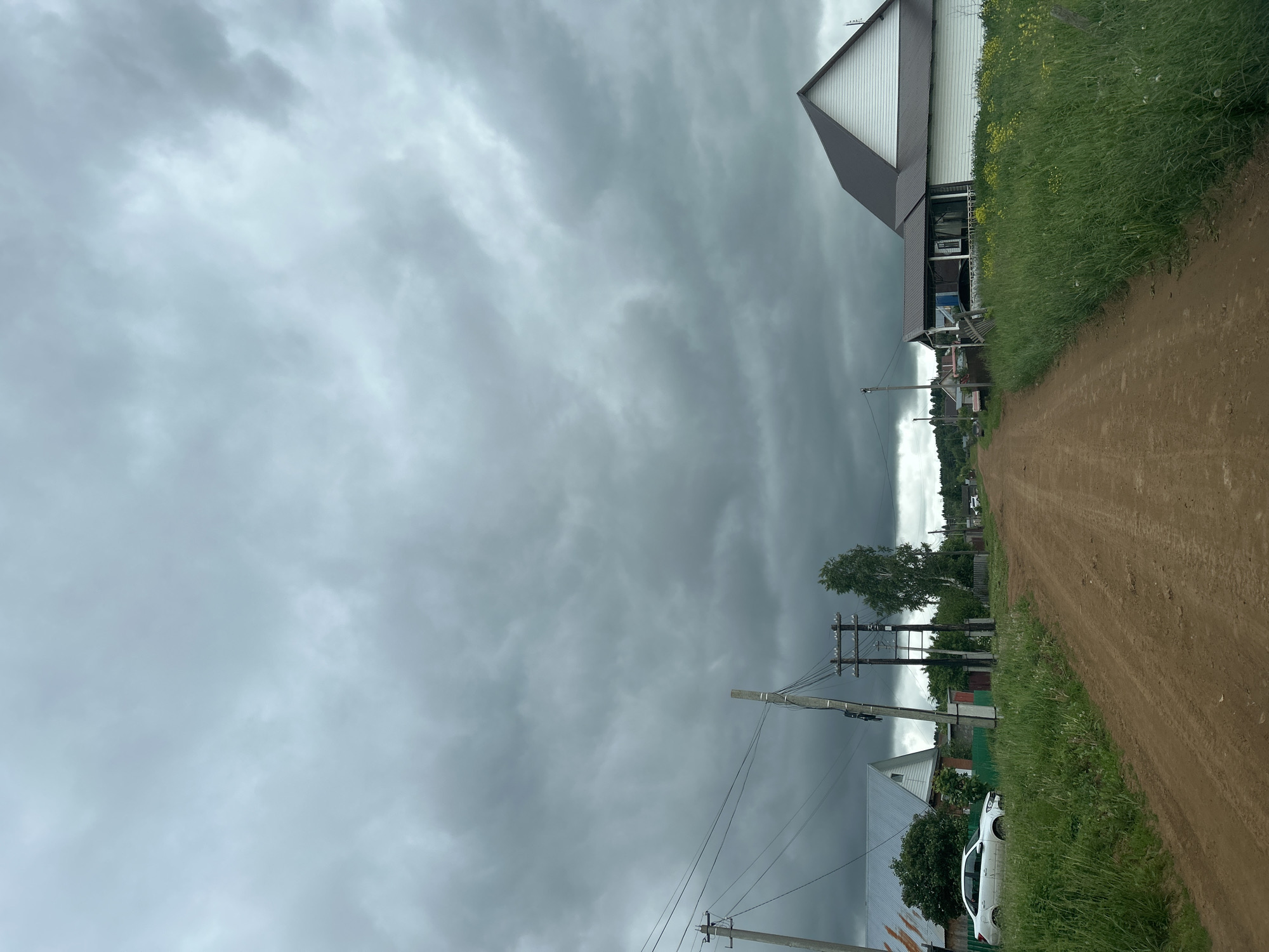 The image depicts a rural village street in Фоки, Russia, under a heavily overcast, dark grey sky. A reddish-brown dirt road, showing tire tracks, extends centrally into the distance, flanked by tall green grass and scattered houses. On the left side, a white sedan is parked near a house with a silver corrugated roof and white siding, partially obscured by green fencing and trees. Several wooden utility poles with power lines line the left side of the road. On the right, a prominent house with a dark brown roof and white siding, featuring a gabled end and an attached covered porch, stands behind a wide verge of tall green grass and yellow wildflowers. Further down the road, additional houses and trees are visible, defining the village landscape. No individuals are present in the scene.