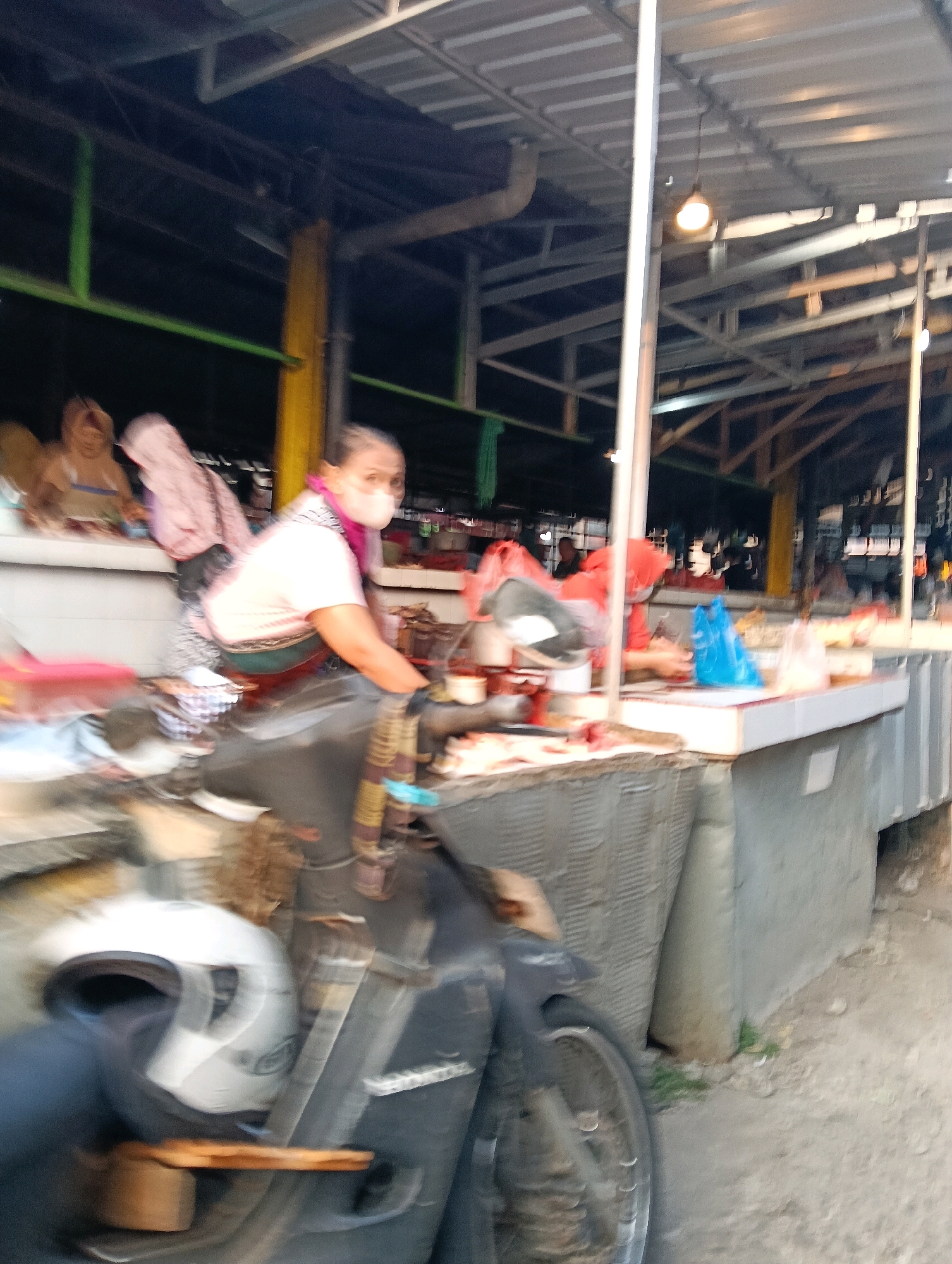 The image depicts a street market scene in Magelang, Indonesia, likely a traditional market given the open-air stalls and the presence of a motorcycle. In the foreground, a portion of a dark-colored motorcycle is visible, with a white helmet positioned on its seat. The market stalls are constructed with a metal roof supported by poles, characteristic of open-air markets.
A woman wearing a face mask and a light pink shirt is the most prominent human subject, positioned behind a market stall laden with various items. She appears to be interacting with the goods on the stall, possibly a vendor. The stall itself has a counter and display of what appears to be food items. Behind her, other individuals are partially visible, engaged in their activities within the market. The environment is bustling, with goods displayed for sale and the general ambiance of a commercial area. The lighting suggests it is daytime, with sunlight filtering through the market structure. The image has a slightly blurred quality, indicating motion or a quick capture.