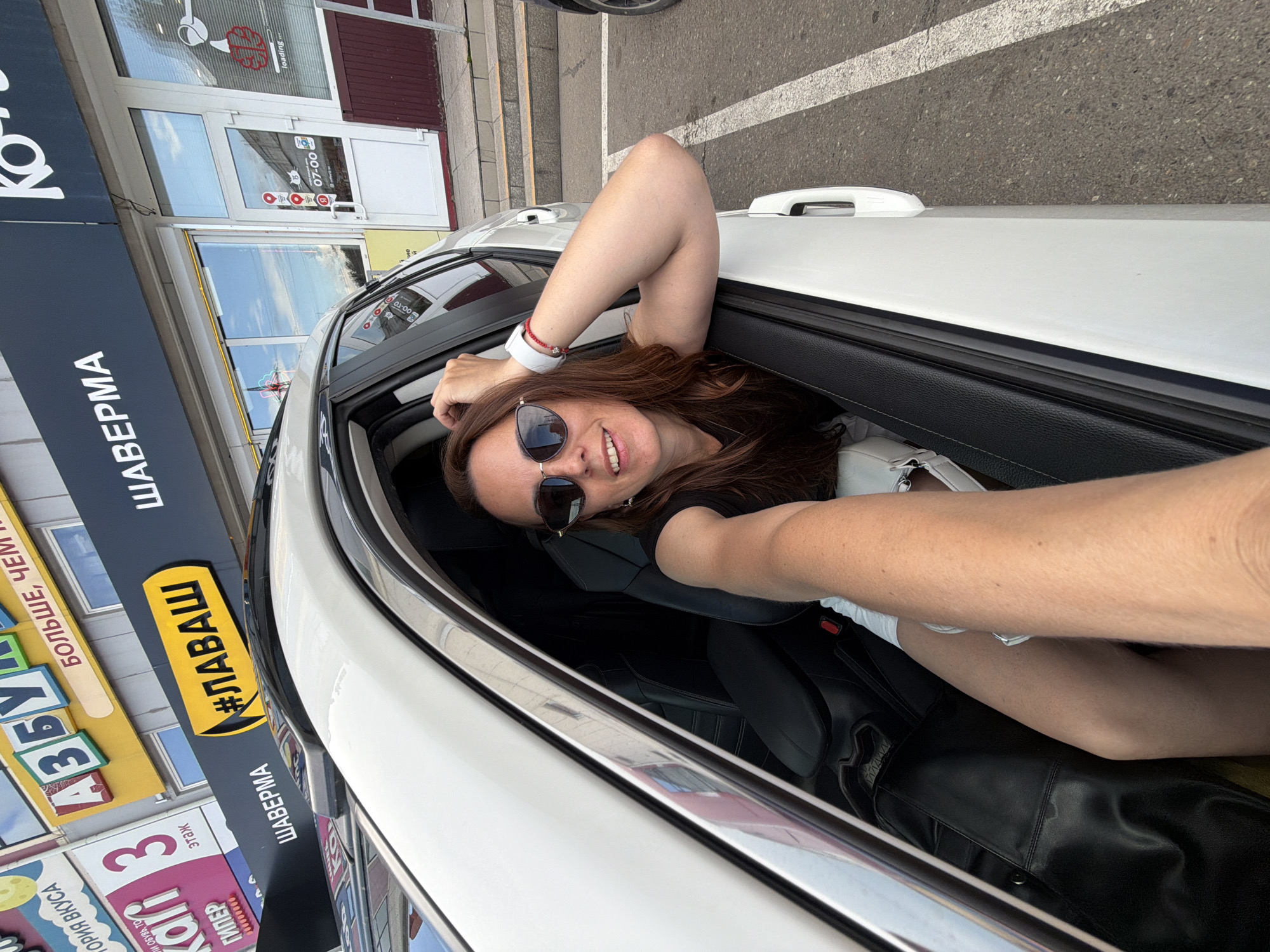 In this vertical, eye-level selfie, a smiling young woman with long brown hair and sunglasses leans out of a white car window. She is wearing a black top and a white bracelet. The car appears to be parked on a street, with a sidewalk and building visible in the background. The building has large windows and signage, with some text visible in Cyrillic script. One sign displays "#ЛАВАШ", suggesting a food establishment. Another sign reads "ШАВЕРМА" which is a Russian dish. The time appears to be daytime, given the bright lighting and reflections in the windows. The woman's pose and smile convey a sense of fun and relaxation. The overall scene suggests a casual outing or a moment captured while traveling.
