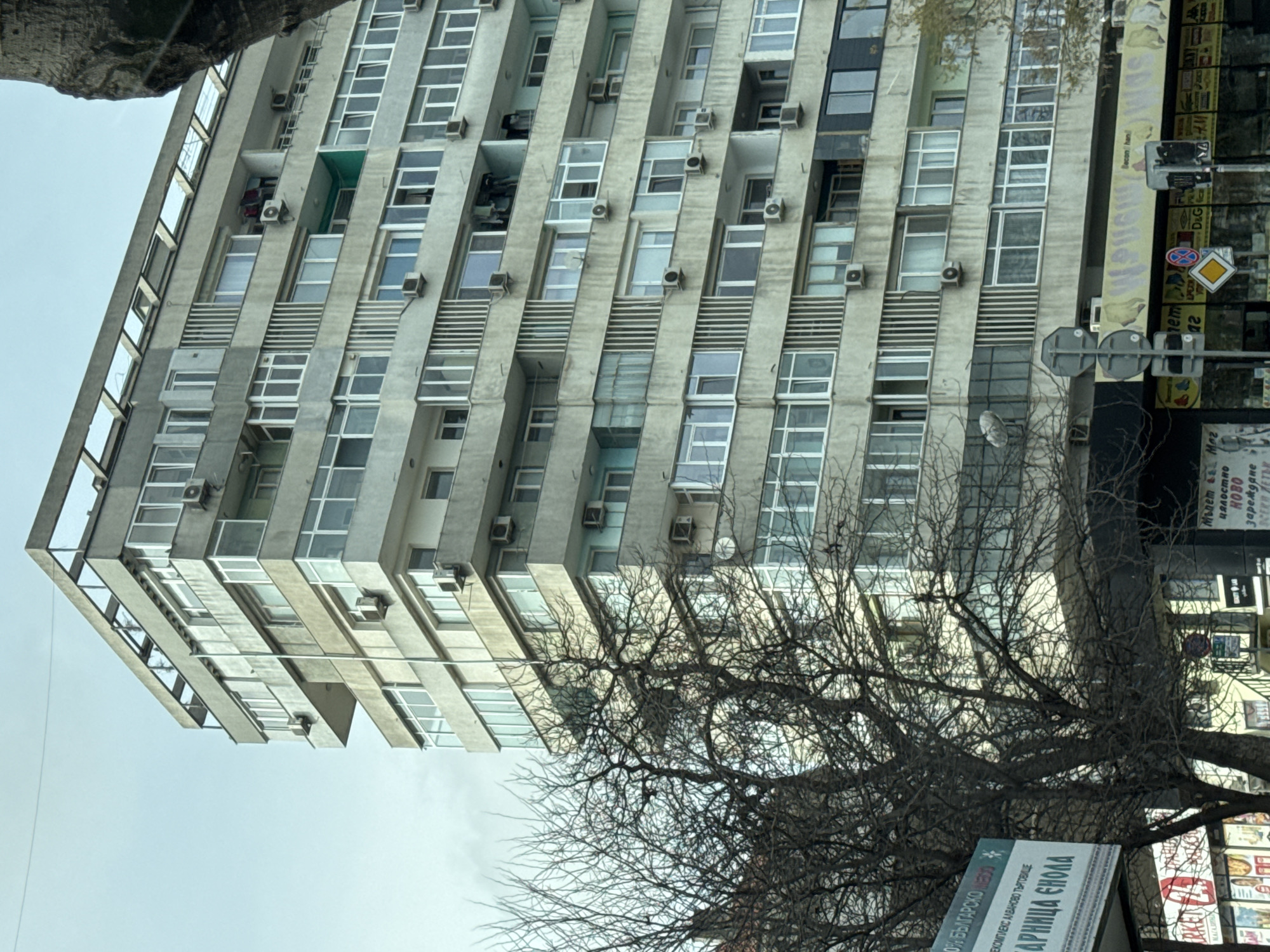 The image shows a tall, grey, concrete apartment building in Varna, Bulgaria, likely constructed in the mid-to-late 20th century. The building has a repetitive facade of stacked rectangular windows and concrete balconies, each potentially housing an air conditioning unit. The building's structural design incorporates horizontal concrete slabs, creating a repetitive pattern of shadowing.
In the foreground, a leafless tree with intricate branching obscures part of the building. The camera angle is tilted, offering a skewed perspective of the building's verticality. A sign for "ARNICA ENOLA" is visible near the base of the tree, potentially indicating the presence of a business or commercial establishment. The context provided by the text on the sign suggests the building may be located near the "ALVANovo TRADE Complex" in Varna. Signs bearing advertisements are visible on the edge of the picture, suggesting a busy intersection. The sky is overcast.