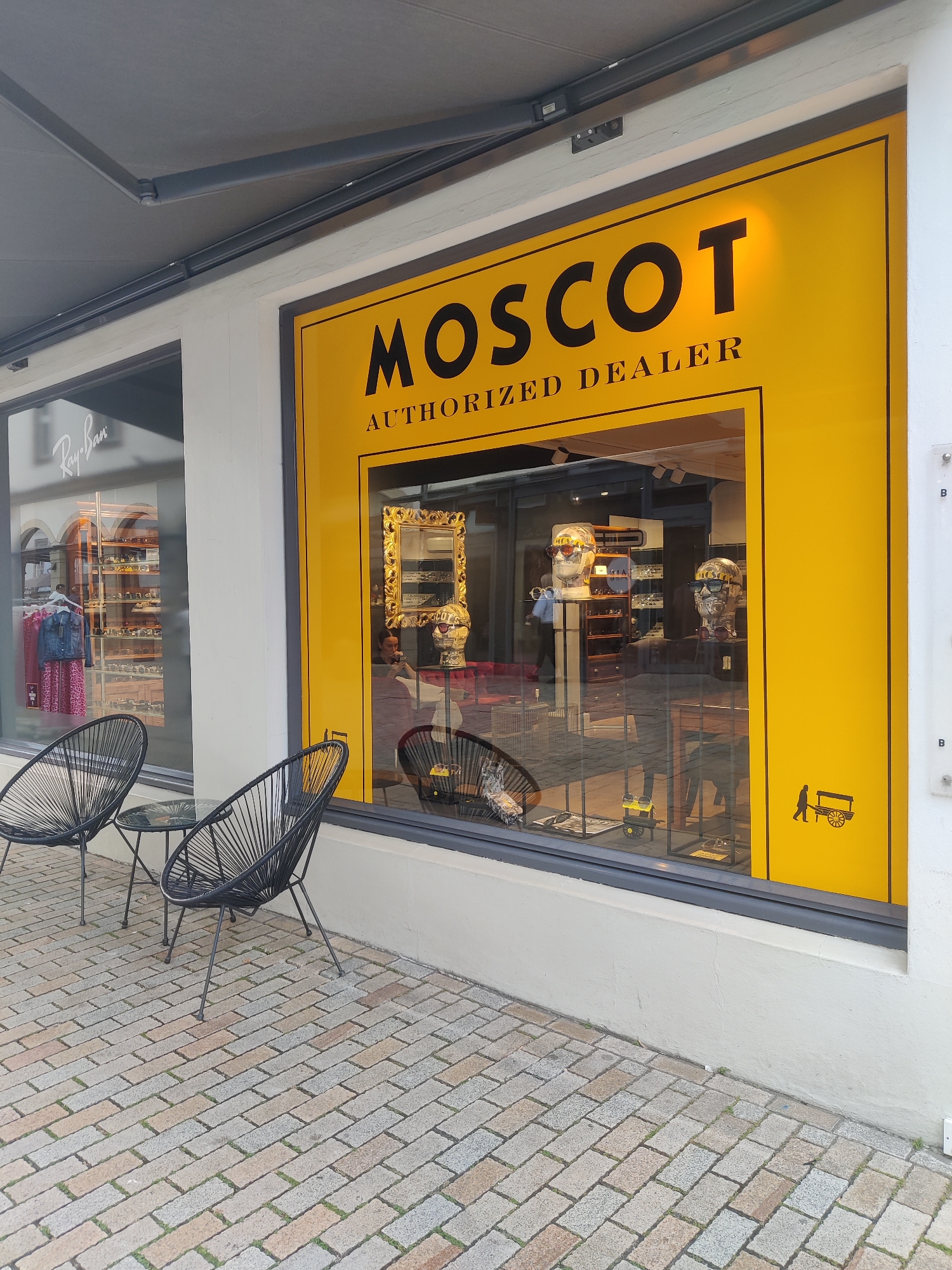 This media file captures the exterior of a MOSCOT authorized dealer store. The prominent feature is the large, bright yellow storefront window with "MOSCOT AUTHORIZED DEALER" clearly printed in black lettering. The window displays an array of eyeglasses on mannequins and shelves, suggesting the store sells optical products.
Outside the store, there are two black, wire-frame Acapulco chairs and a small round table, set on a cobblestone sidewalk. The lighting suggests it is daytime, possibly late morning or early afternoon. The sky is not visible, so the weather cannot be determined.
To the left of the MOSCOT store, a portion of another shop's window is visible, featuring a "Ray-Ban" logo and displays of sunglasses and clothing. This indicates a commercial area, likely a shopping street, in Bielefeld, Germany. The overall impression is of a bright, modern retail environment.
Visible text includes "MOSCOT AUTHORIZED DEALER" and "Ray-Ban." There are also small silhouette graphics of a person pushing a cart on the yellow part of the window. Reflections in the store window show some interior details and a person inside.