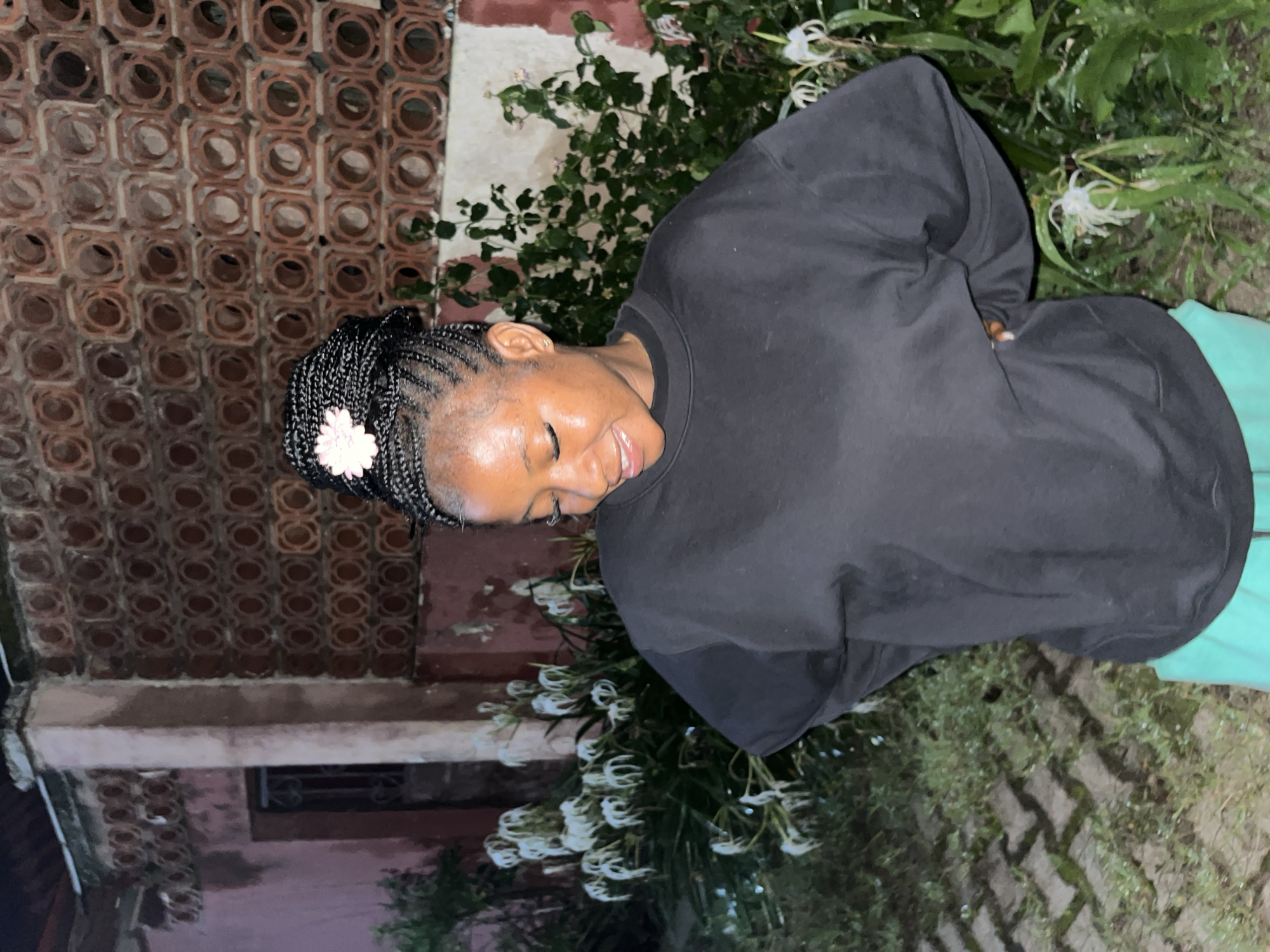 A young Black woman with braided hair styled in a bun, adorned with a pink flower, stands outside at night in what appears to be a residential area in Jos, Nigeria. She is wearing a black crewneck sweatshirt and light green shorts. Her hands are placed on her hips as she looks down and smiles. The background features a textured brick wall with circular openings, a weathered pink pillar, and lush green foliage with white flowers. The ground is paved with interlocking bricks interspersed with grass.