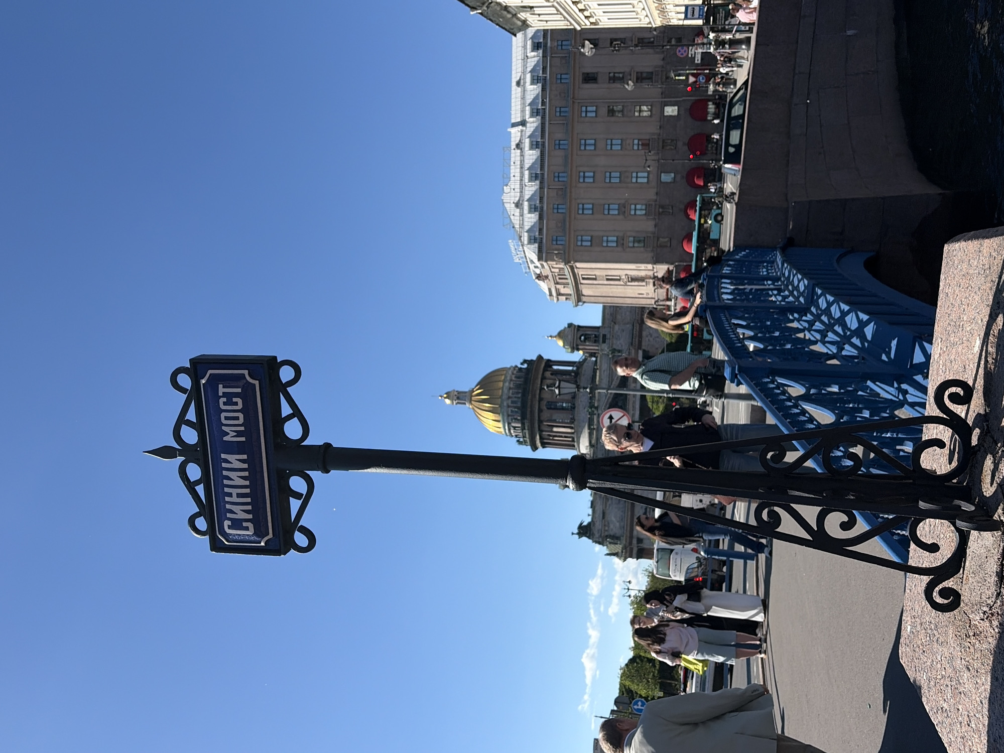 The image shows a street sign in Saint Petersburg, Russia, that reads "СИНИИ МОСТ" (Blue Bridge) in Cyrillic letters. The sign is mounted on an ornate black lamppost. In the background, the golden dome of Saint Isaac's Cathedral is visible. Several people are walking on the street and a blue bridge crosses a canal. The weather is sunny with a clear blue sky. The scene appears to be a busy urban environment with historical architecture.