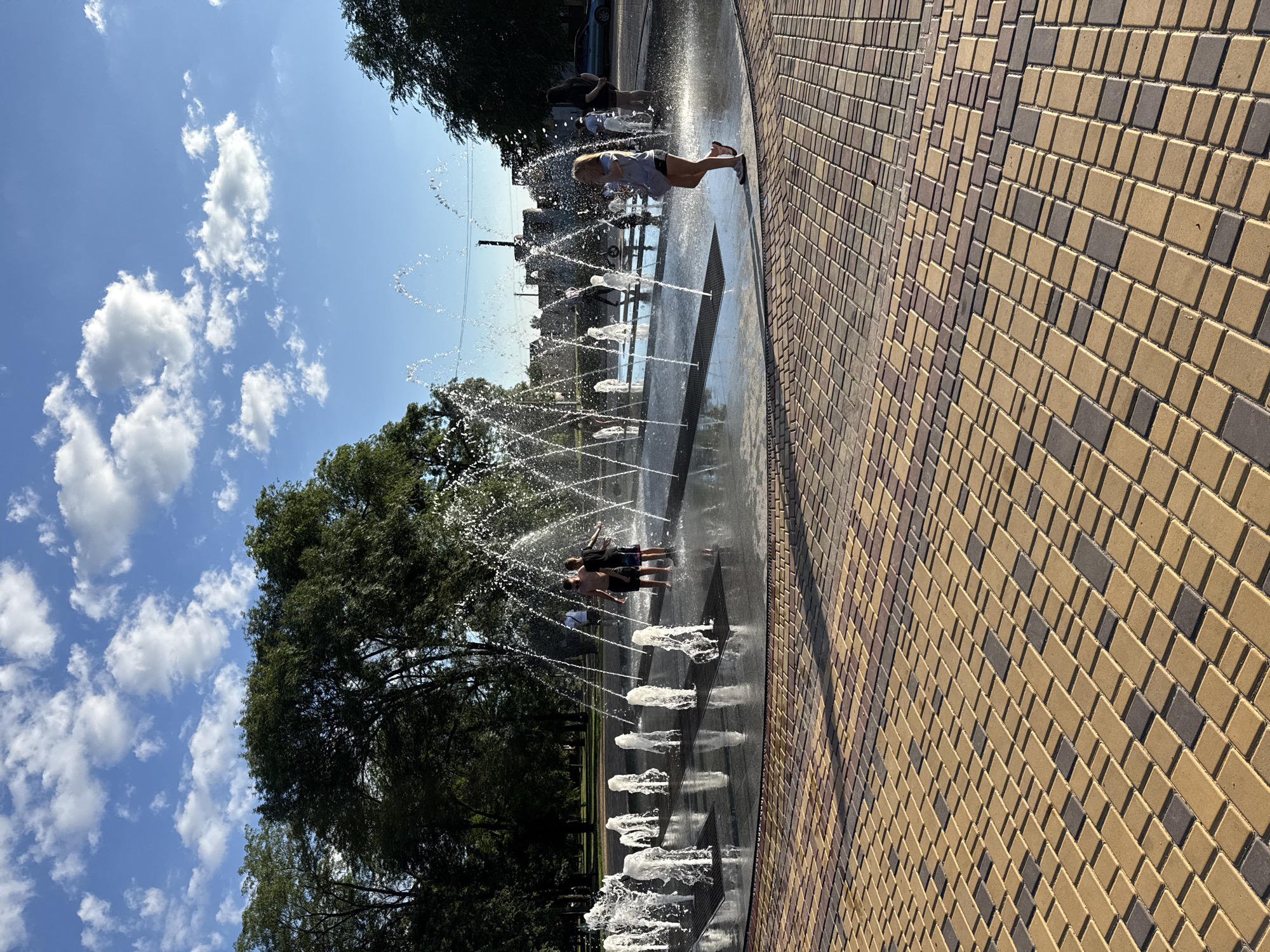 In this image, a group of people are enjoying a public fountain on a sunny day. The fountain is designed with numerous jets of water spraying upwards from the ground, creating a playful and refreshing atmosphere.
The subjects are primarily children and young adults, scattered throughout the fountain area. One young girl in the foreground, with blonde hair, is captured mid-action, leaning back as water sprays around her. Further back, a group of three individuals, two boys and a girl, are standing together, also interacting with the water jets. Other figures can be seen in the background, suggesting a lively and populated space.
The setting appears to be an urban park or public square, with lush green trees lining the background. The ground around the fountain is paved with a patterned brickwork, in shades of yellow and brown, which adds to the visual appeal of the scene. The sky above is a vibrant blue, dotted with fluffy white clouds, indicating fair weather. The sunlight casts distinct shadows, suggesting it is mid-day. The overall emotion conveyed is one of joy and carefree fun, as people cool off from the heat of the day. There is no visible text in the image.