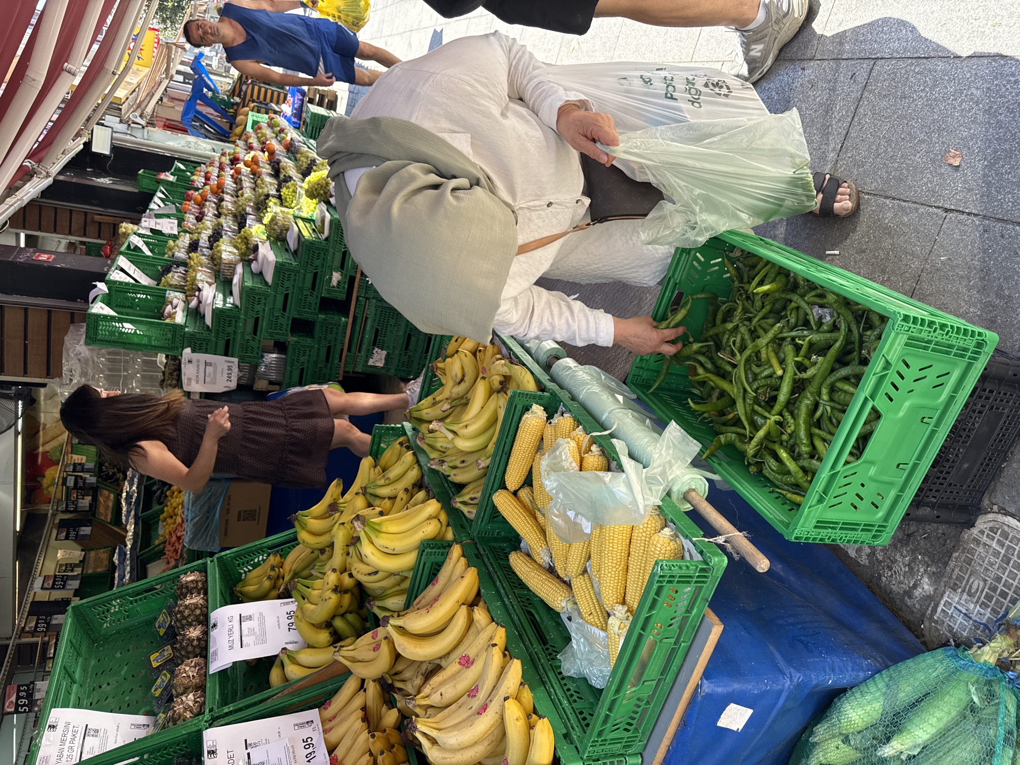 An outdoor market scene is captured in Kadıköy, Turkey, showcasing a vibrant display of fresh produce. A woman, dressed in a white long-sleeved shirt and a light green headscarf, is seen from the waist down, selecting green chili peppers from a bright green plastic crate. She holds a transparent plastic bag in her right hand. To her left, a crate filled with corn on the cob is partially visible, with some corn in clear plastic bags. In the background, a variety of fruits, including bananas and what appear to be pineapples, are arranged in green crates. A woman in a dark dress walks by in the middle ground, and a man in a blue tank top and shorts stands to the right, holding a yellow plastic bag. The market stalls are set up on a paved sidewalk under a striped awning.