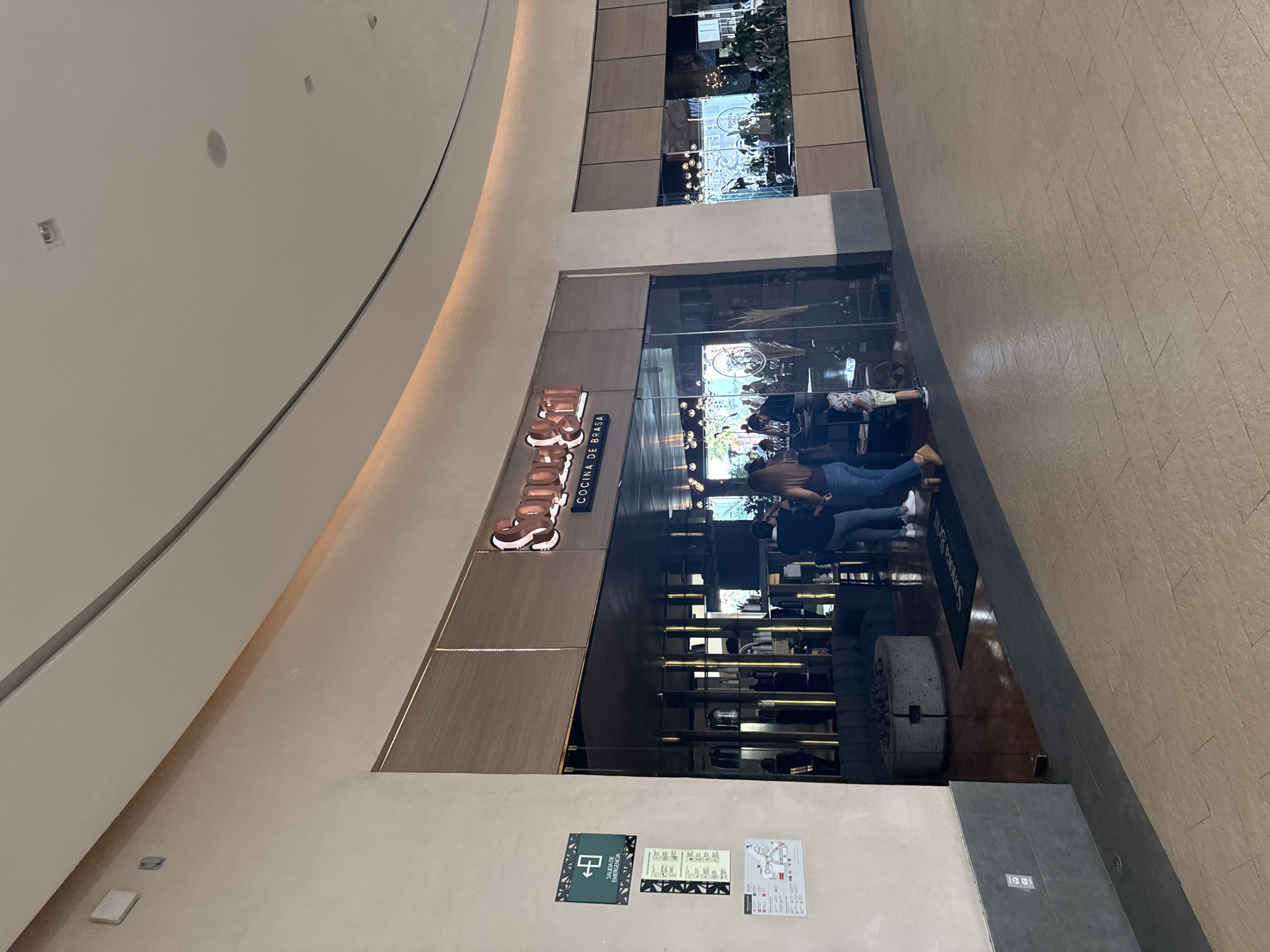 The image captures the Sonora Grill restaurant in the city of Puebla, Mexico. The facade of the restaurant, featuring the brand name "Sonora Grill" and the words "COCINA DE BRASA" in Spanish, is visible. Inside the restaurant, patrons are visible. Signage with safety information is affixed to the wall near the entrance. The surrounding environment appears to be an indoor space.