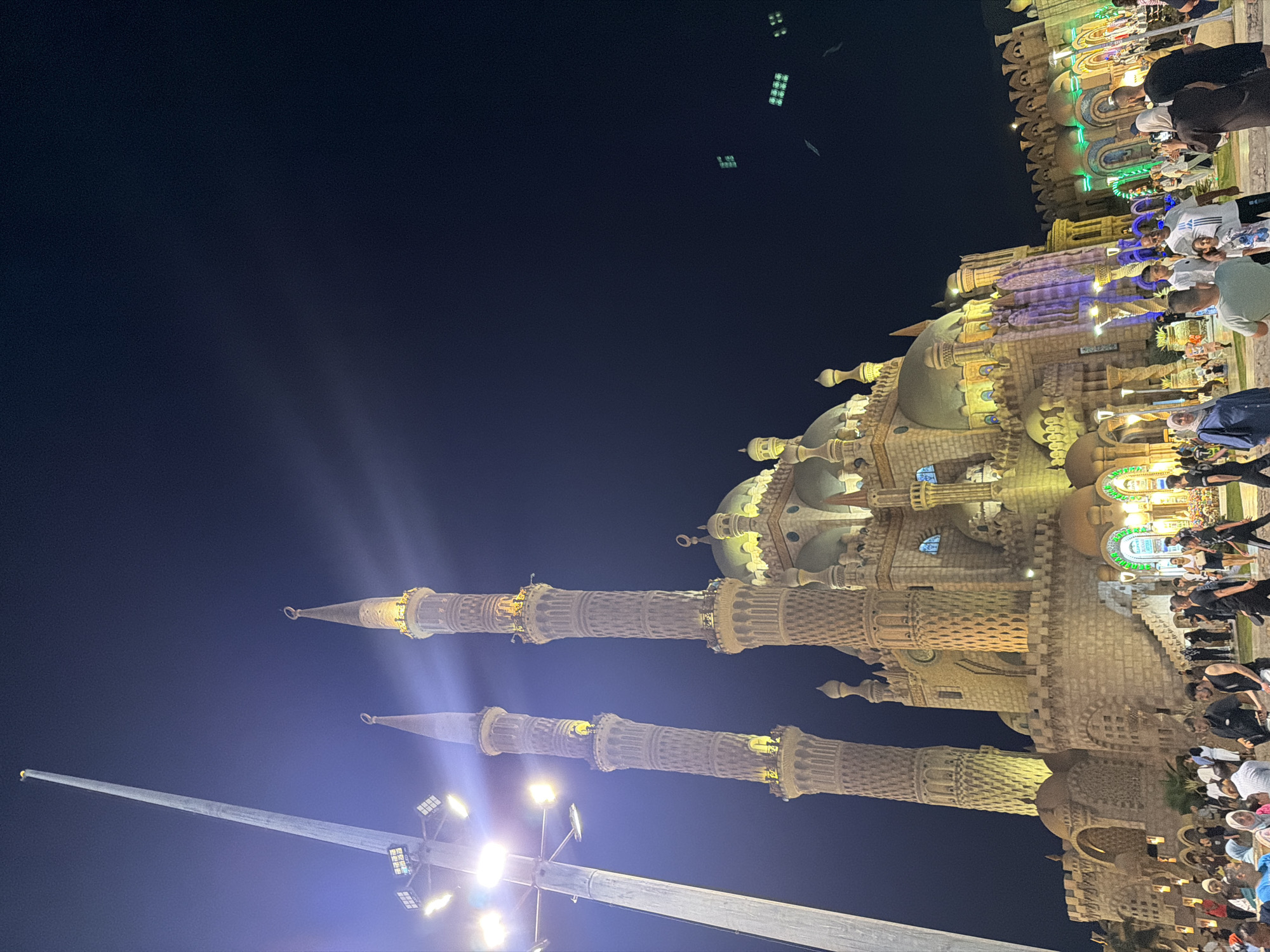 The image captures the Al-Sahaba Mosque in Sharm El Sheikh, Egypt, at night. The mosque's distinctive architecture, characterized by its intricate stonework, domes, and prominent minarets, is illuminated by artificial lighting, casting a warm glow against the dark sky. Two tall minarets, intricately carved and topped with crescents, flank the central structure. A tall light pole with multiple spotlights stands to the left, its beams cutting through the night. In front of the mosque, a crowd of people is gathered, some walking, others standing and observing. The people are diverse in age and attire, with many dressed in casual clothing. The atmosphere appears to be lively, suggesting a popular gathering spot. The surrounding area is decorated with lights, adding to the festive ambiance of the nighttime scene.