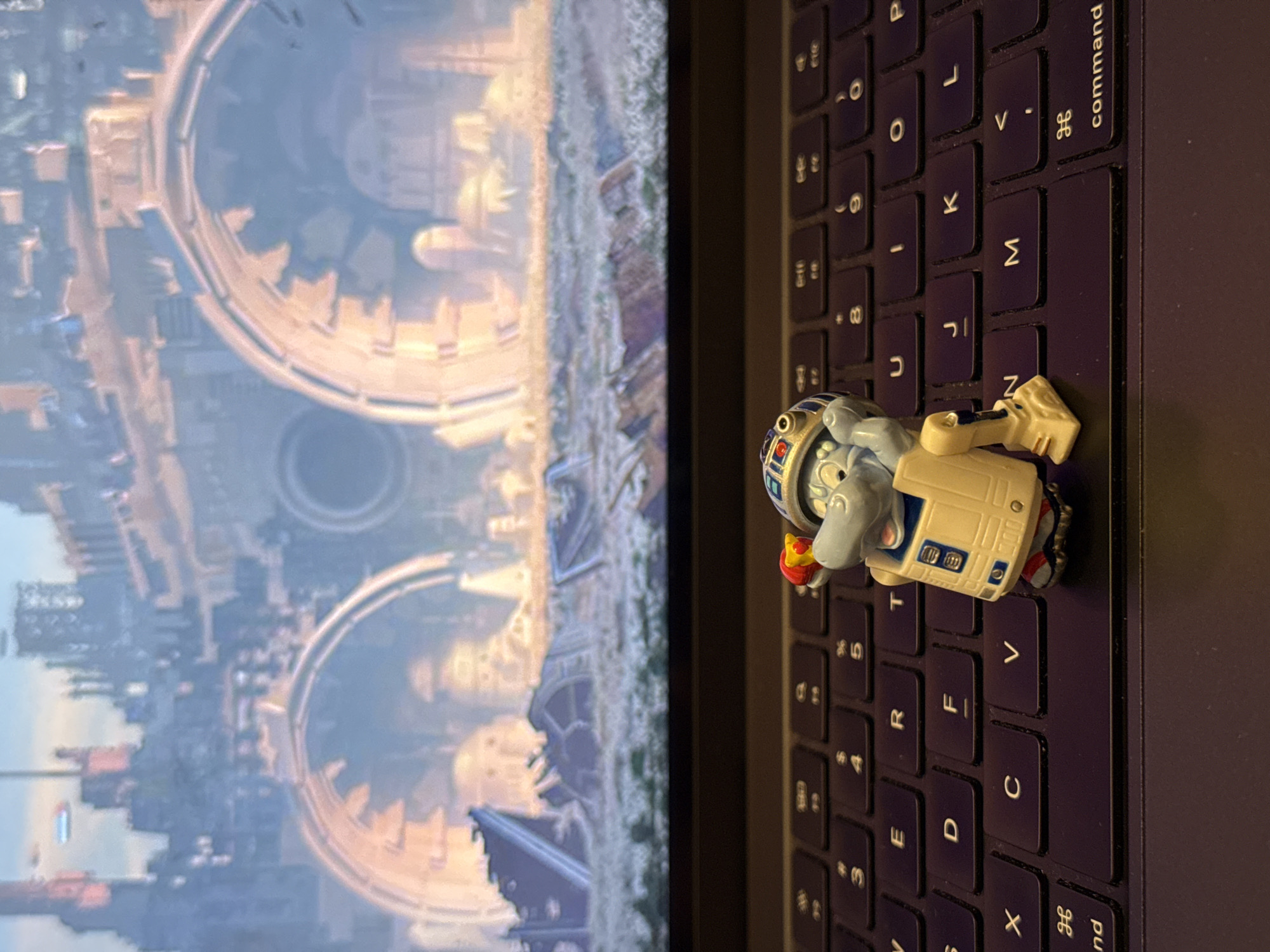 The image depicts a screen showing a location that appears to be in Moscow, Russia. The structures visible on the screen resemble those found in the city. A toy-like figure rests on the keyboard of the laptop, positioned near the center of the image. The keyboard is primarily black, with white characters on the keys. The background is dominated by the screen, which is illuminated and showing a cityscape with distinct architectural elements. The laptop itself is partially visible, with the screen taking up a significant portion of the frame. The lighting in the image appears to be ambient, casting soft shadows around the objects. The focus is primarily on the toy and the keyboard, with the background providing context to the setting.