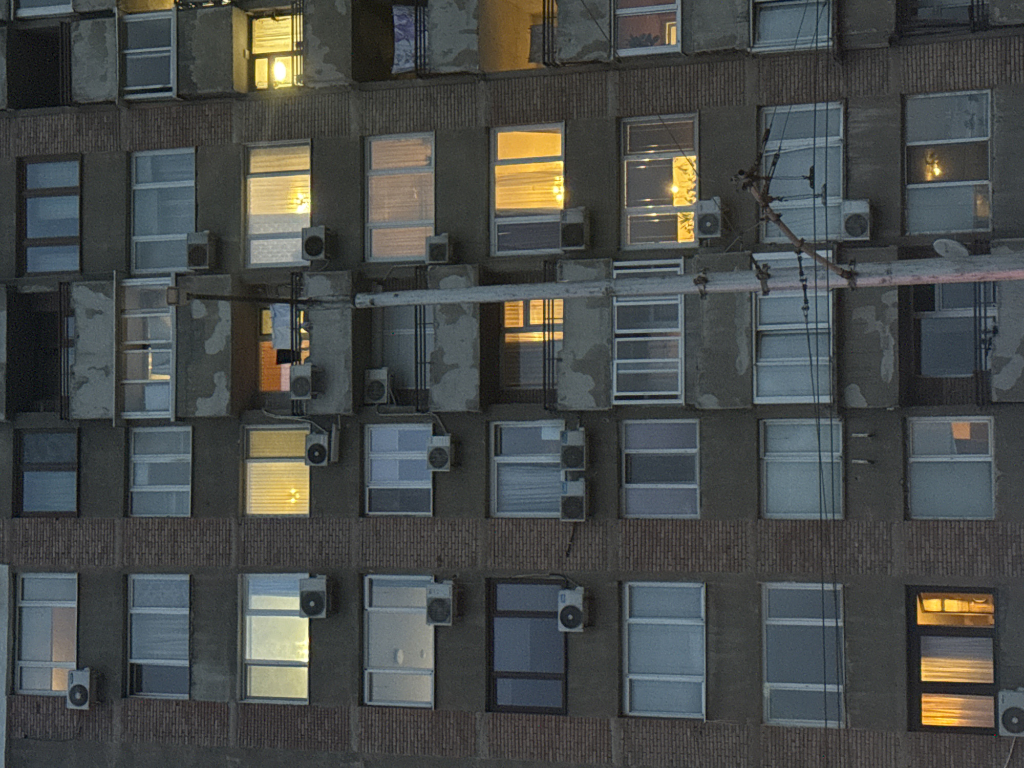 An exterior shot of a residential apartment building in Sofia, Bulgaria, shows multiple levels of windows, some with lights on, some with blinds, and some dark. Each window has a dark gray frame and an air conditioning unit installed underneath. The exterior of the building has horizontal bands of brickwork separated by gray concrete. A utility pole runs across the building.