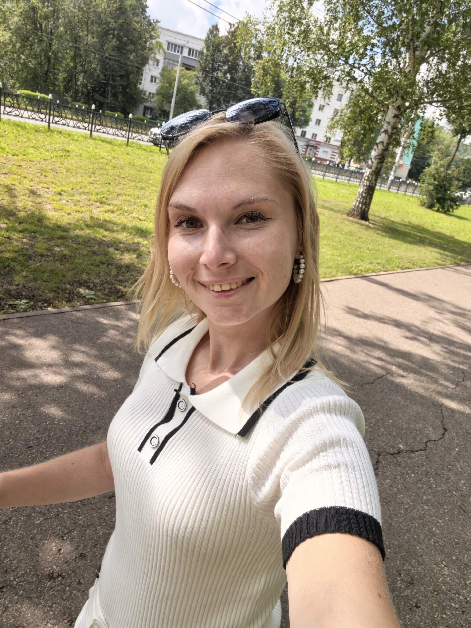 The image is a close-up, selfie-style photograph of a Caucasian woman with blonde hair and fair skin. She is outdoors in a park-like setting, possibly in the city of Sterlitamak, Russia. The woman is smiling and wearing a white, ribbed polo shirt with black trim and buttons. She has on hoop earrings with pearl-like beads and sunglasses perched on top of her head. The background features a paved walkway, green grass, trees, and a building. The lighting suggests it is daytime with sunlight casting shadows. The image is focused on the woman's face and upper body.