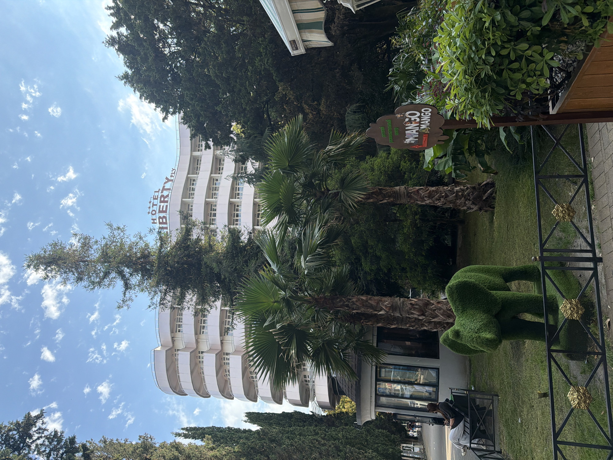 The image shows a sunny day in Sochi, Russia, with a view of the Hotel Liberty Fly building. The hotel is white with curved balconies and a sign at the top. In front of the hotel, there are palm trees and other greenery. A topiary elephant made of grass stands in the foreground. A woman is sitting on a bench to the left, looking at her phone. A sign with "MANGO & MANGO" written on it is visible to the right. The environment appears to be a park or hotel grounds.