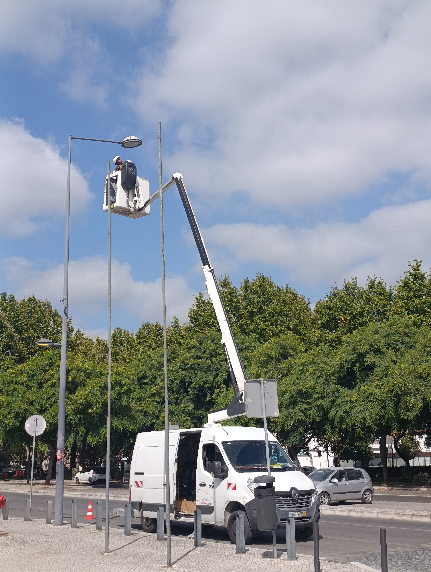 A street maintenance worker is positioned in a bucket lift, suspended from an articulated arm attached to a white van. The worker is wearing a hard hat and appears to be inspecting or repairing a streetlamp mounted on a tall pole. The van is parked on the side of a road, with a sidewalk made of patterned paving stones in the foreground. Behind the van and sidewalk, a dense line of green trees forms a backdrop. In the distance, several cars are visible parked or driving on the road. The sky above is blue with scattered white clouds. The scene is set in a city environment, likely an urban or suburban area. The license plate on the van reads "19-ZG-75". The location is the Avenida Luísa Todi in Setúbal, Portugal.