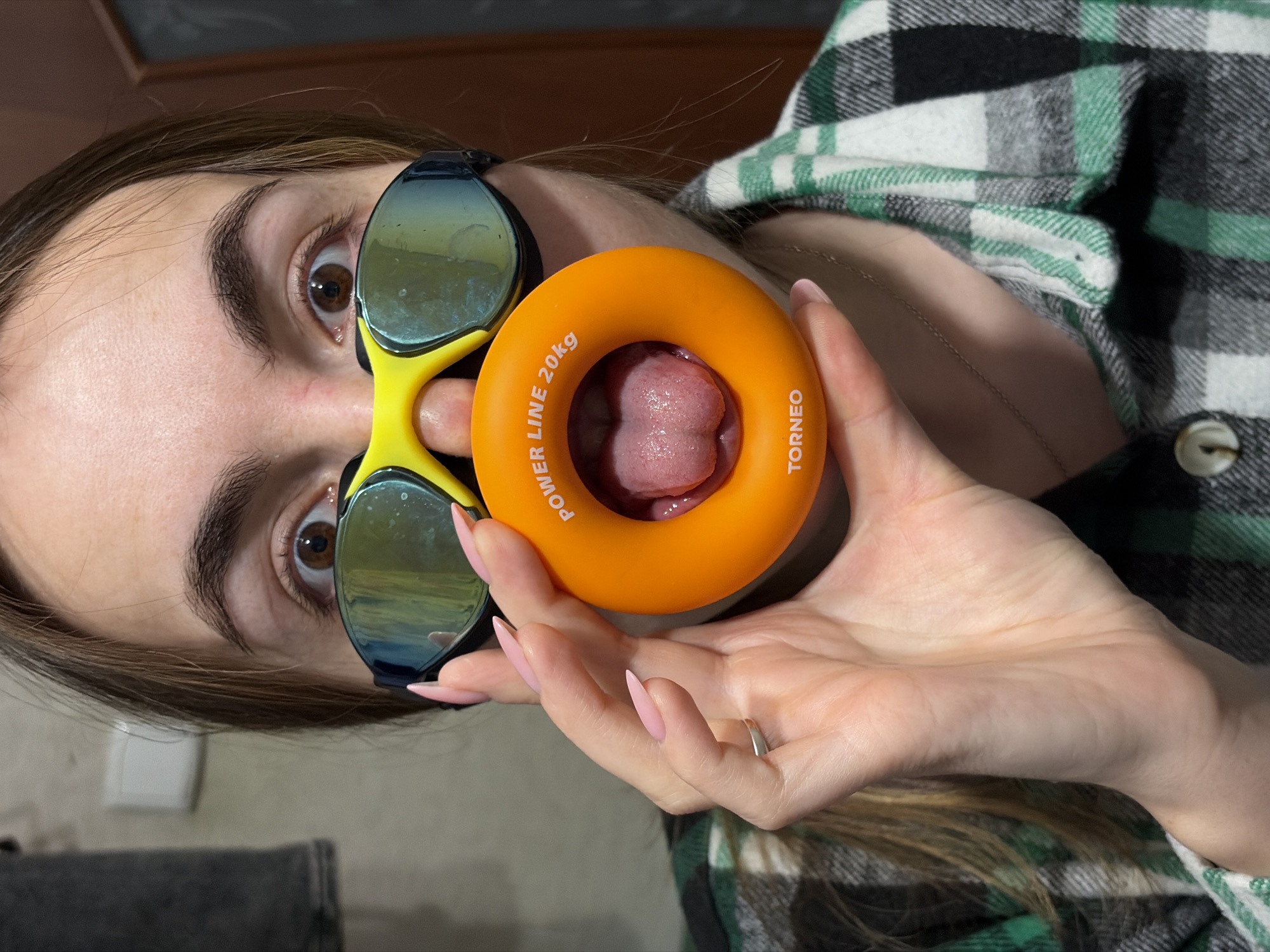 A person wearing swim goggles is holding a "TORNEO" hand grip with a tongue protruding through the hole. The hand grip is orange and labeled "POWER LINE 20kg". The person's nails are painted pink. The person is wearing a green, black, and white plaid shirt. The location is possibly Izhevsk, Russia (implied). The image is rotated counter-clockwise.