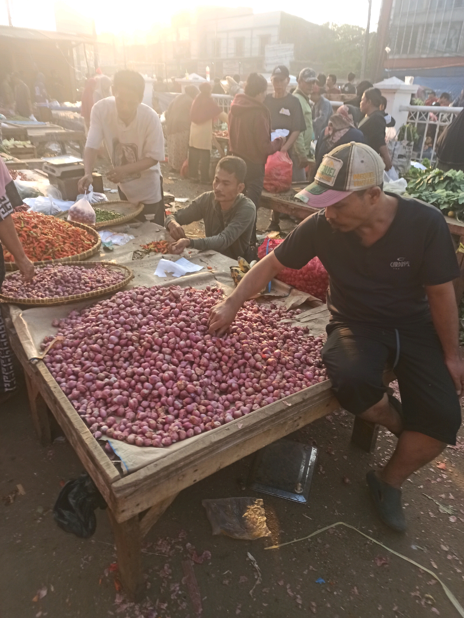 The image depicts a bustling outdoor market in Cikarang, Indonesia, likely in the early morning given the soft, hazy sunlight. A man in a black t-shirt and shorts is seated on the edge of a wooden table laden with a large pile of red onions. He is reaching towards the onions with his right hand. To his left, another man in a gray long-sleeved shirt is also interacting with produce on the table, which includes smaller piles of red chili peppers and what appears to be green onions or shallots. Behind them, a variety of people are visible, some engaged in transactions, others browsing or standing. Stalls and market structures are present in the background, suggesting a typical Indonesian market environment. The ground is dirt and appears somewhat scattered with debris. The overall impression is one of a vibrant, active marketplace.