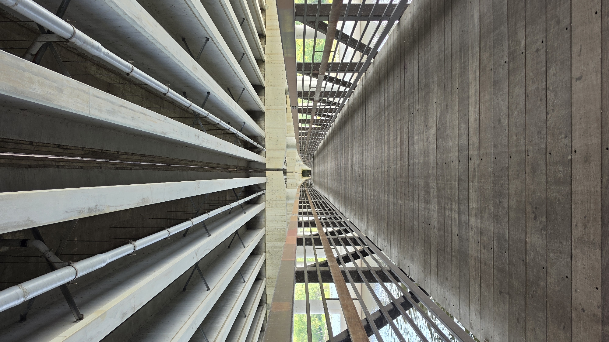 The image depicts the underside of an overpass and a pedestrian bridge leading towards it in Elgin, United States of America. The viewpoint is from the pedestrian bridge, looking down its length towards the overpass. The bridge is constructed of weathered wooden planks, laid horizontally and running the length of the walkway. Metal railings, with a dark brown or black frame and horizontal wooden slats, line both sides of the pedestrian bridge.
Above the pedestrian bridge, the underside of the overpass is visible. It is composed of parallel concrete beams, running longitudinally and supporting the structure above. These beams are light gray and have a somewhat weathered appearance. Running between the concrete beams are several large, silver pipes, likely for utilities, which are secured by metal clamps. Darker, thinner pipes and wiring are also visible, attached to the beams and pipes above.
The environment surrounding the pedestrian bridge suggests it is located near water, as a body of water with ripples is visible to the left of the bridge. Green foliage and trees are seen in the background through the gaps in the railing, indicating an outdoor setting with natural elements. The lighting suggests it is daytime, with shadows cast by the overpass structure. The scene is devoid of any people.