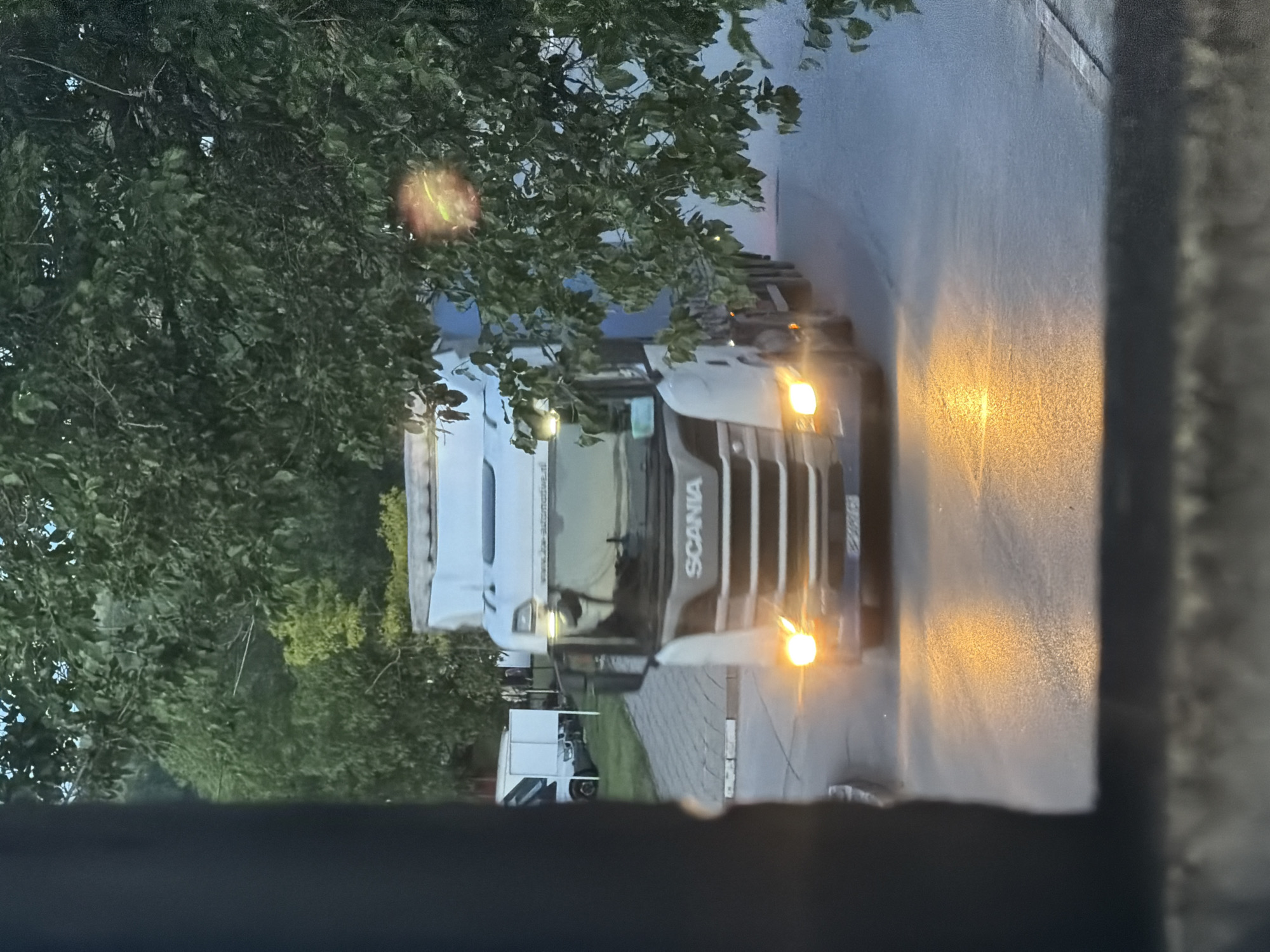 The image, oriented vertically, captures a white and dark grey Scania semi-truck driving on a paved road, likely in Iskar, Bulgaria. The truck's front is prominent, with "SCANIA" clearly visible on its grille. Its headlights and amber marker lights are illuminated, casting bright light on the road surface and a textured, light-colored wall or building on the right side of the frame. This illumination, combined with the overall dimness, suggests the time of day is either dusk, dawn, or an overcast day.
Behind the truck's cab, part of a white trailer is visible. Faint text on the side of the trailer appears to be a web address: "www.u2g-automotive.de". Another smaller white vehicle, possibly a van or light truck, can be seen in the background on the left, partially obscured by trees.
The left side and foreground of the image are filled with lush green foliage from numerous trees, some branches hanging low and partially obscuring the view. The road surface appears dark and somewhat reflective. No people are visible in the scene. The truck's license plate is present but unreadable due to the angle and lighting. The setting suggests an industrial or roadside environment bordered by greenery.