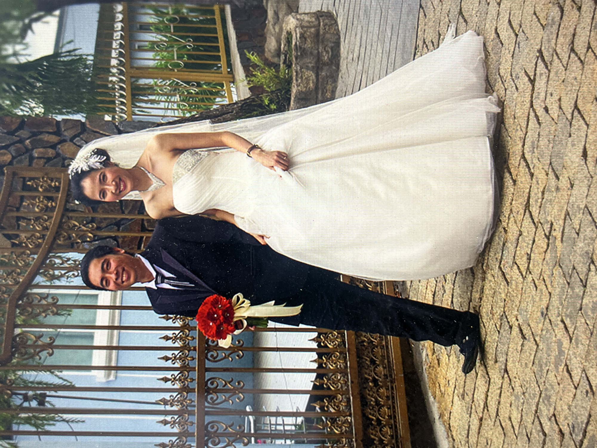 A wedding photo taken outdoors features a bride and groom. The bride wears a white strapless wedding gown with a veil and a floral headpiece. She has her hand on the groom's shoulder. The groom wears a black suit with a white shirt and black tie, holding a boutonniere of red flowers. They stand in front of an ornate gold metal gate. Behind the gate, a light blue building is visible. The background includes foliage, a stone structure, and a stone pathway or sidewalk with a textured surface. Based on the geographic details, this photo may have been taken in the city of Bac Nha Trang Ward, Vietnam.