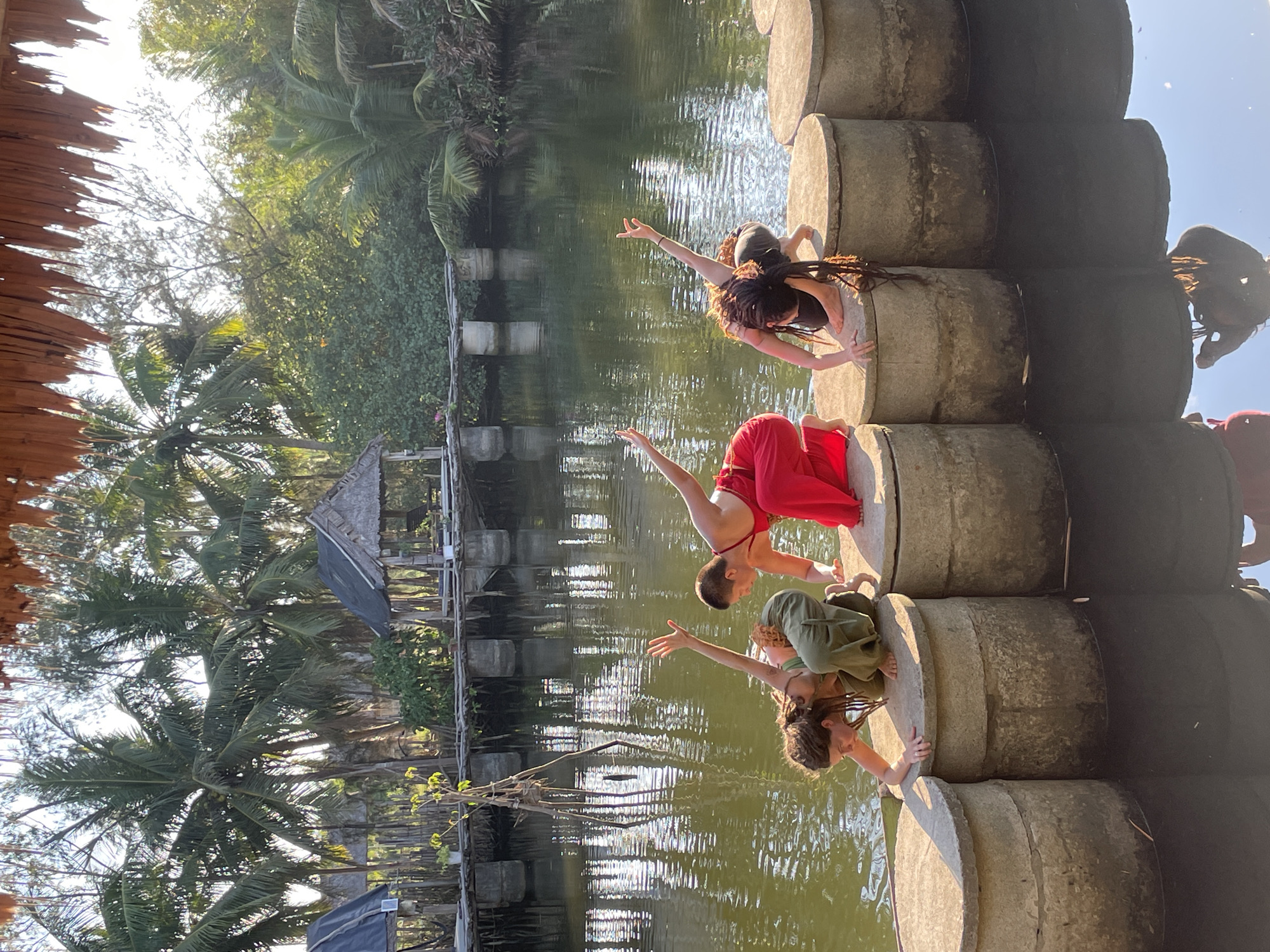 Three individuals are positioned on a series of cylindrical concrete structures in a body of water, possibly a pond or a river, in Đà Nẵng, Vietnam. The individuals are wearing varying attire: one is in red, another in green, and the third in black. They appear to be engaging in a form of dance or expressive movement, with arms extended. The water reflects the scene. Lush vegetation and palm trees surround the area, and there are structures in the background. A thatched roof can be seen in the foreground.