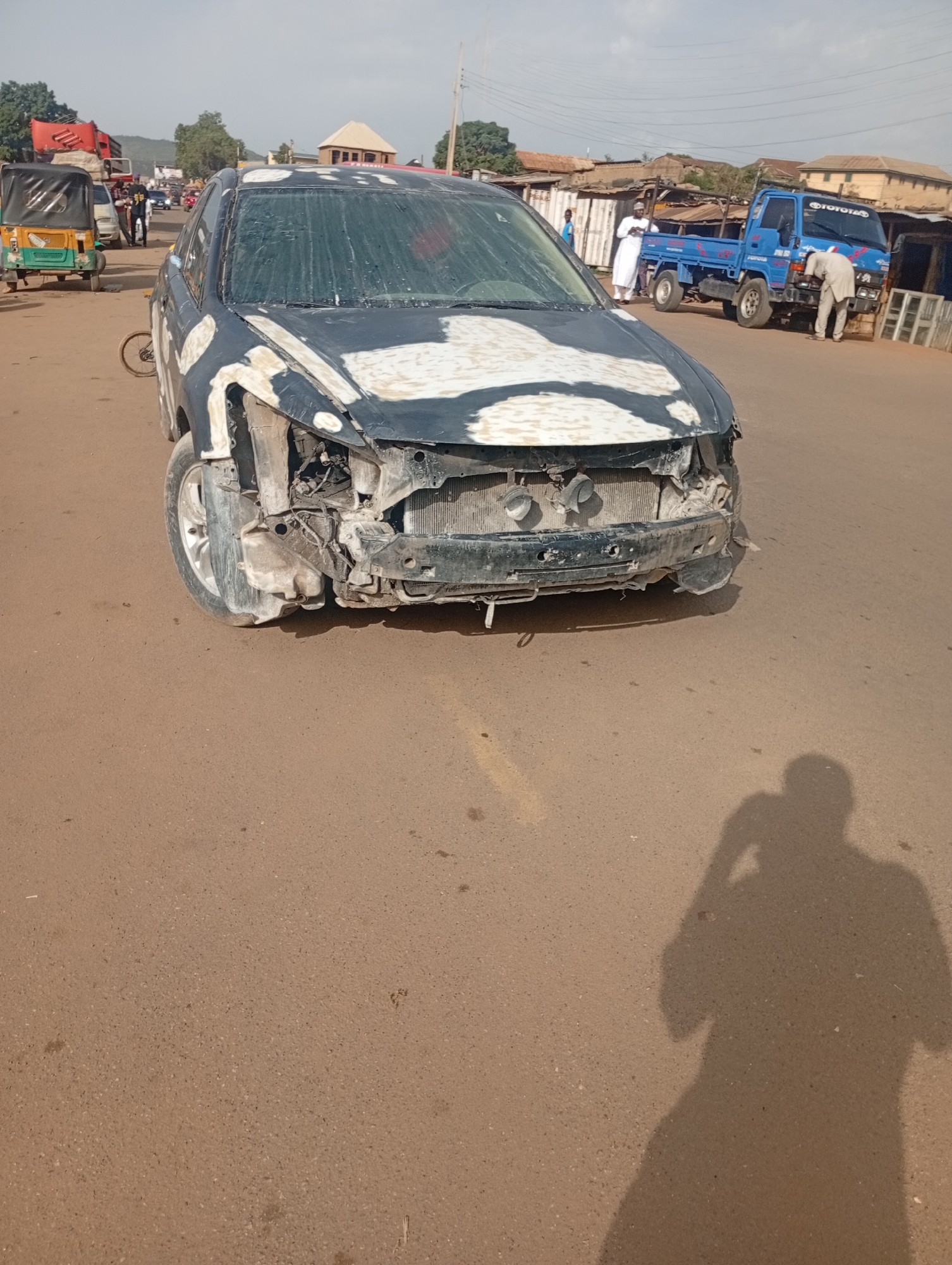 A damaged black car sits in the middle of a dirt road in Rafin Par, Nigeria. The car's front end is heavily damaged, with the hood and bumper missing, exposing the engine and radiator. The windshield is covered in white paint splatters, and the car's paint is chipped and peeling. A tri-colored auto-rickshaw is parked to the left of the car, and a blue truck is parked to the right. Several people are visible in the background, including a person in a white robe standing next to the truck and another person in a tan outfit inspecting the truck. The surroundings appear to be a semi-urban area with buildings and market stalls visible in the distance. The road is unpaved and dusty, with tire tracks and debris scattered on the surface. A shadow of a person taking a photograph is cast on the road in the foreground.