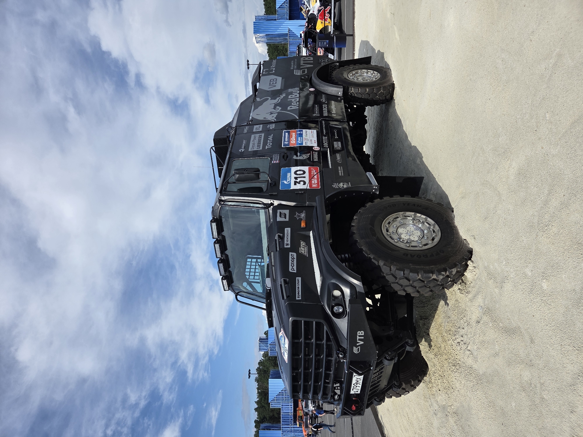 This is a low-angle, full-shot photograph taken outdoors on a bright, cloudy day, likely at an event or exhibition in Yekaterinburg, Russia. The primary subject is a large, matte black, off-road truck, which appears to be a racing or support vehicle. The truck has numerous sponsor logos and decals visible on its sides and front, including "Red Bull," "GAZPROM," "KAMAZ," "VTB," "LIEBHERR," and "TOTAL." A prominent number "310" is displayed on the side. The truck is positioned on a sandy surface with tire tracks visible. In the background, there are large blue shipping containers, a line of trees, and other vehicles, some of which also appear to be racing vehicles with bright livery. A few individuals are visible in the distance, walking. The sky is partly cloudy with streaks of white cirrus clouds against a blue background. The overall scene suggests a motorsport event or a display related to rally racing.