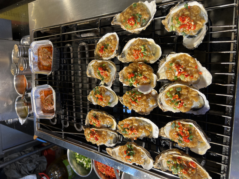 This image presents a detailed view of grilled seafood preparation within a commercial food environment in Yuzhong District, China. The central focus is a metal grate grill, actively cooking approximately 19 to 20 oysters arranged in their half-shells. Each oyster features a translucent vermicelli noodle base topped with a thick, light brown minced garlic or savory sauce, further garnished with finely chopped red chilies and green scallions.
In the upper right portion of the grill, two rectangular aluminum foil containers hold a reddish, saucy dish, possibly a meat or vegetable preparation. Behind these containers, a series of kitchen items are visible, including several small, cylindrical metal condiment shakers, a small stainless steel bowl with a reddish liquid or sauce, and another small stainless steel bowl containing an oily liquid.
To the upper left of the grill, a large, clear plastic bottle with a green label is partially visible. Adjacent to it are two stainless steel bowls: one filled with finely chopped green scallions and the other with finely chopped red chilies. The overall setting implies a food stall or kitchen area characterized by stainless steel surfaces and essential cooking ingredients. No individuals are present within the frame.