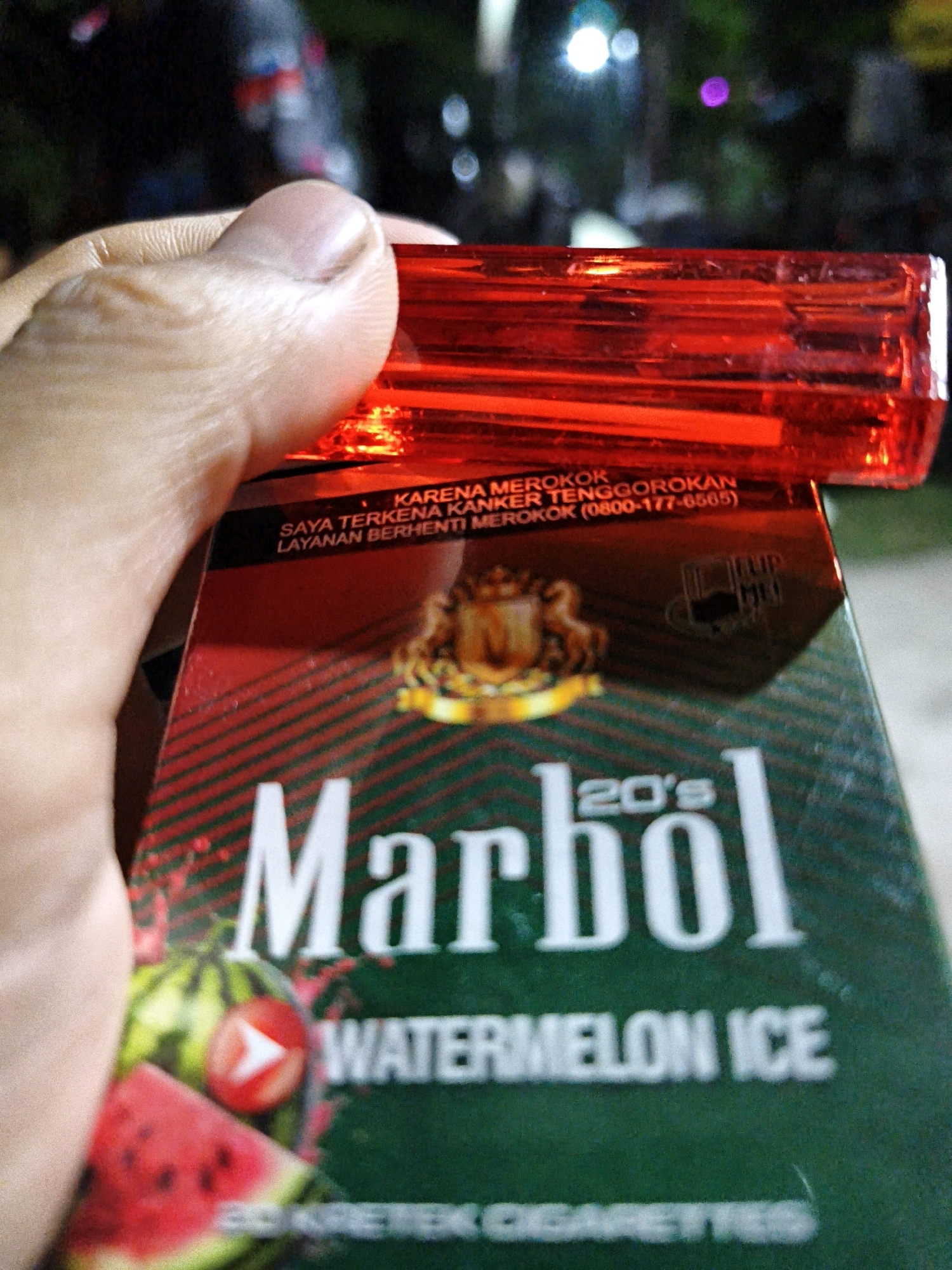 An outdoor, medium shot captures a person's hand holding a red lighter over a pack of "Marbol 20's Watermelon Ice" kretek cigarettes. The cigarette pack features images of watermelon slices and a logo. Text on the pack indicates a smoking cessation hotline. The background is blurred, showing a dark, outdoor environment with some indistinct lights, suggesting it may be nighttime. The scene is likely set in Pengkok, Indonesia, a location known for its agricultural areas and rural lifestyle.