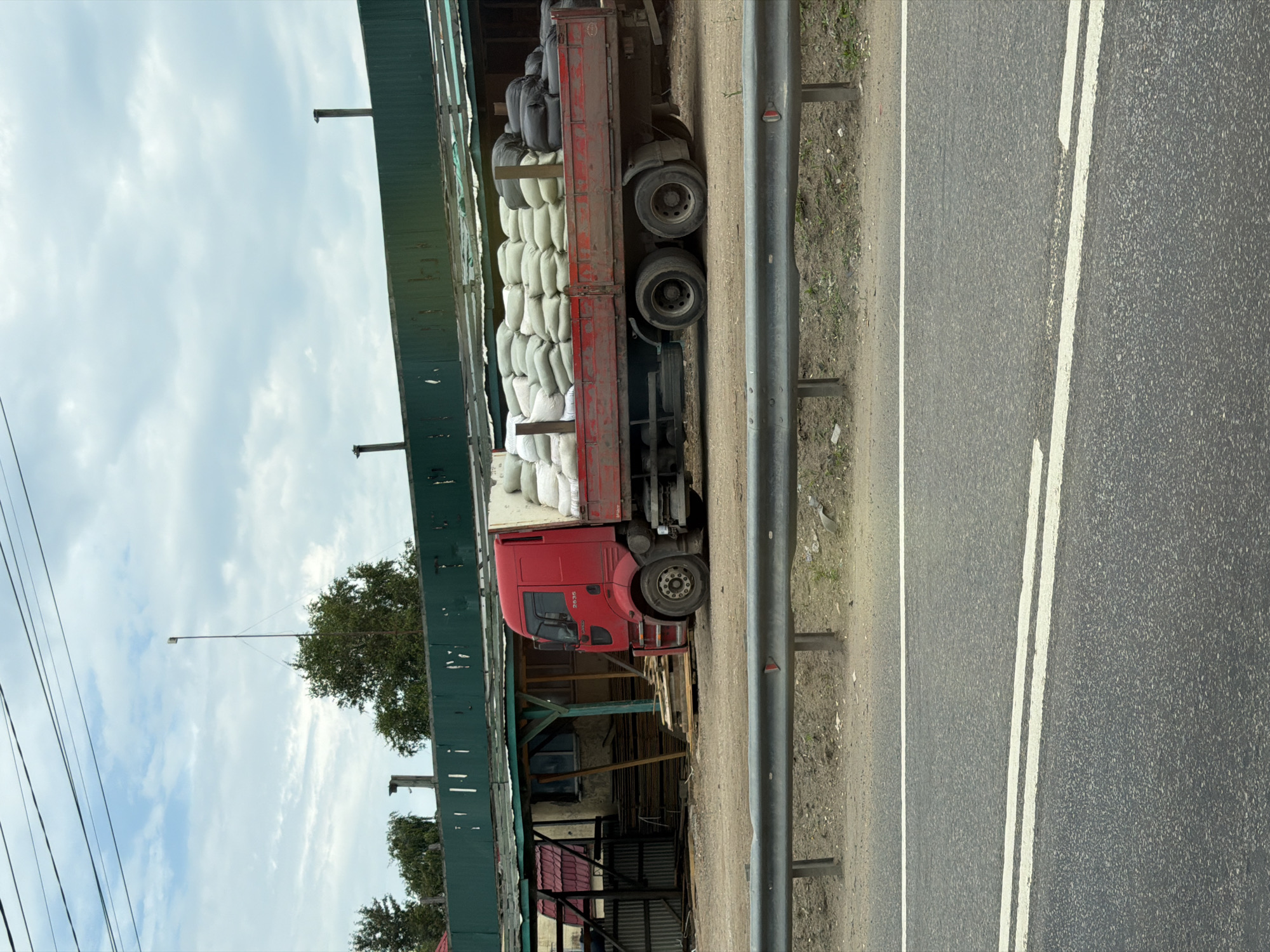 A red flatbed truck is parked on a dirt lot in front of a low-rise building with a green corrugated metal roof. The truck bed is filled with rows of white and black sacks. The building appears to be a workshop or storage area with wooden beams and corrugated metal siding. In the background, there are trees and a cloudy sky. A metal guardrail runs along the edge of the road in the foreground. The scene is likely located in Radumlya, Russia, given the visual context.