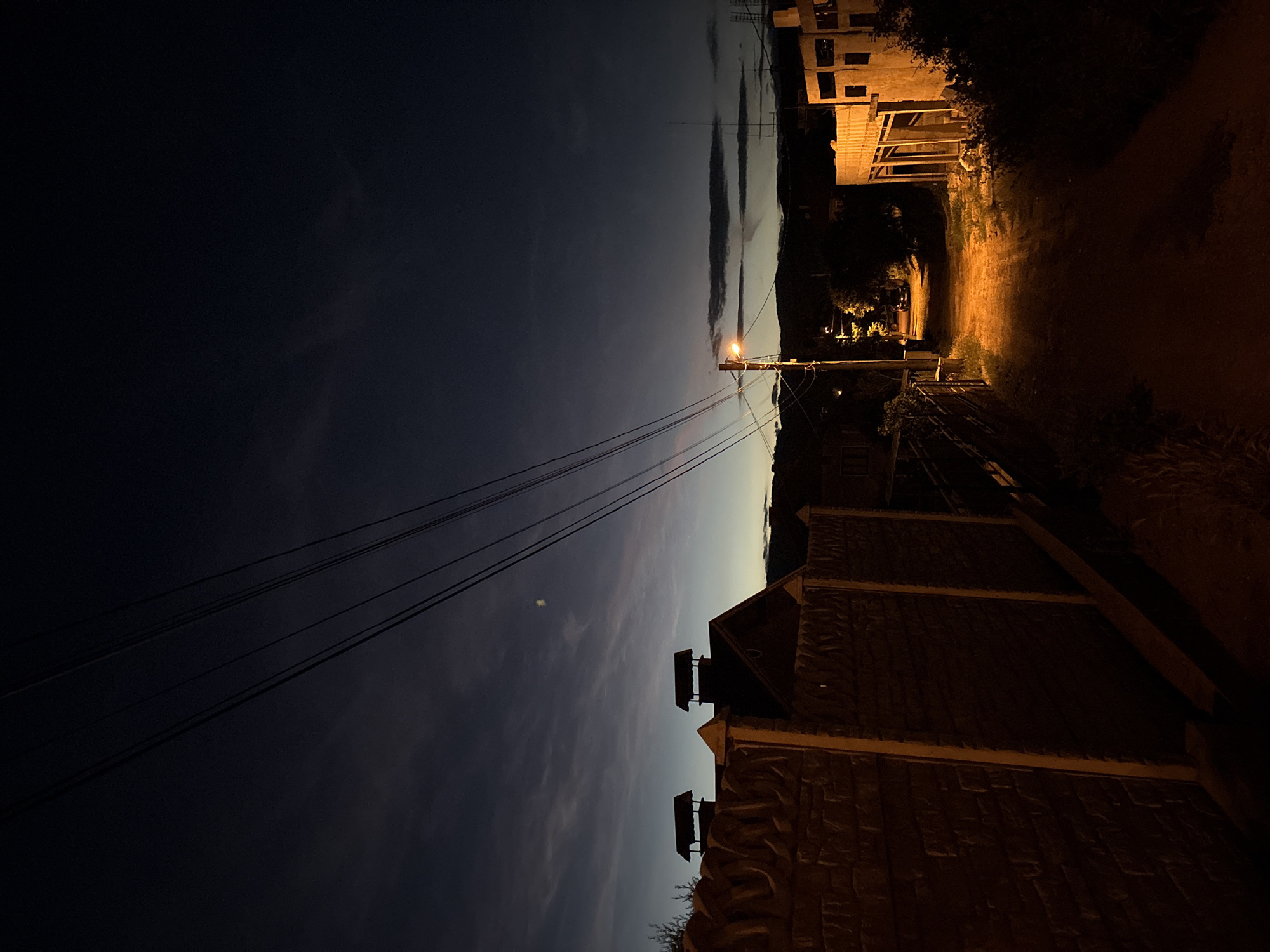 An outdoor, full shot captures a dirt road at dusk in the village of Podgumer, Bulgaria. A tall, textured concrete wall runs along the left side of the road. Two brick chimneys with dark, sloped roofs rise above the wall. Overhead, four parallel black wires extend diagonally across the dark blue sky, which is streaked with faint, wispy clouds. A single streetlamp, casting an orange glow, is positioned on a wooden pole on the right side of the road. Beyond the streetlamp, trees and houses are visible in the dim light. The road surface is a mix of dirt and grass.