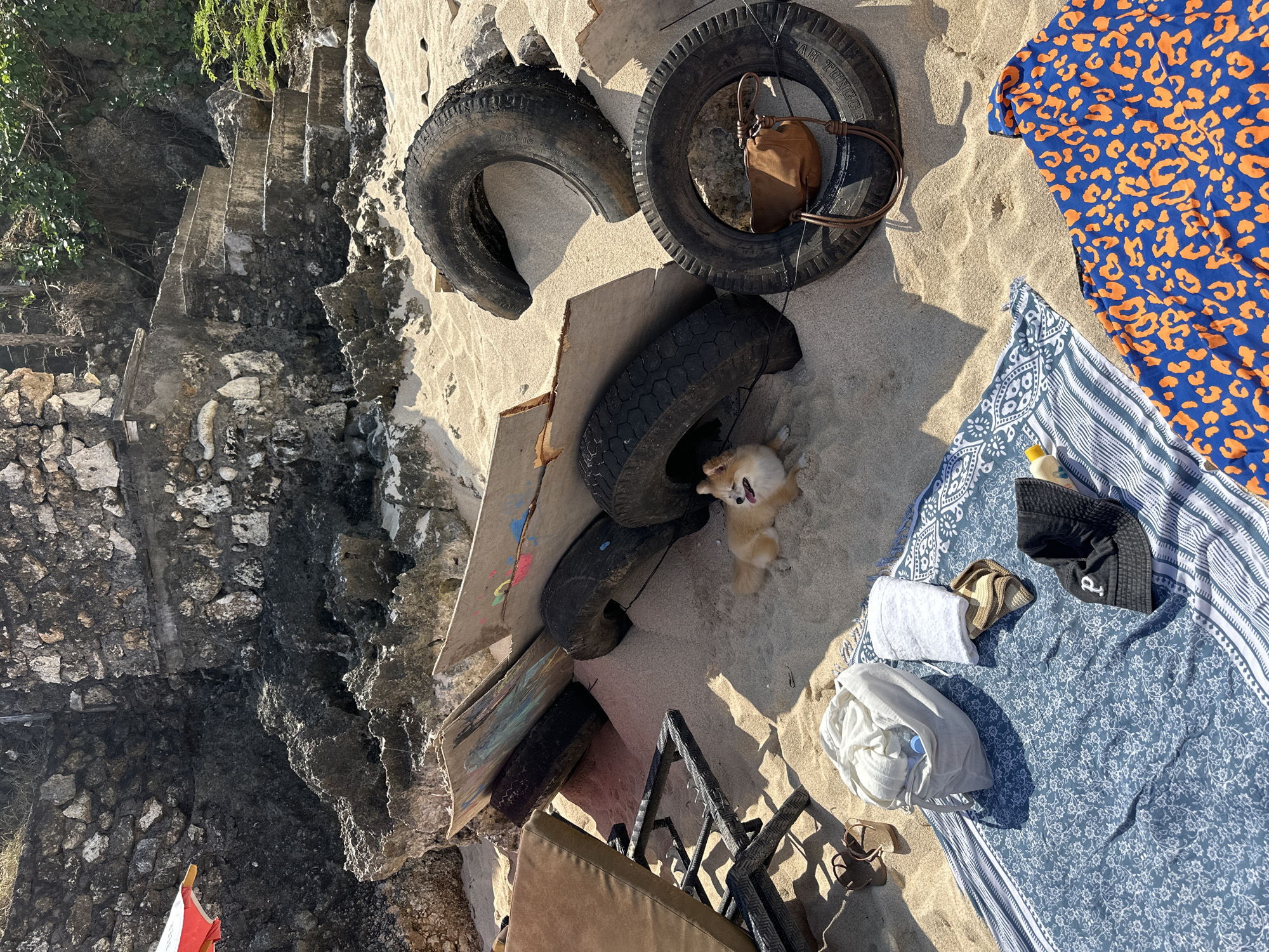 A small, light-colored dog with its tongue out is lying on the sandy beach near Kuta Selatan, Indonesia, amongst several old tires and some weathered wooden boards. To the left, a portion of a lounge chair and a pair of sandals are visible. In the foreground, several towels, a drawstring bag, a bucket hat, and a small yellow bottle are arranged on a blue patterned blanket. To the right of the tires, a brown purse is hanging from one of them. In the background, a rocky cliff face with some greenery and stone steps leads up to a grassy area. Sunlight casts shadows across the sand.
