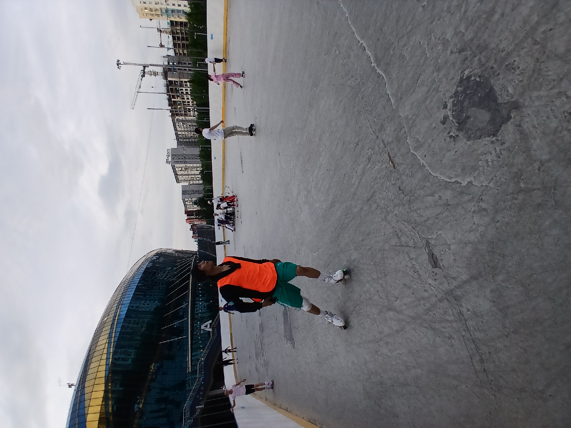 In Unknown, Unknown, a man in an orange vest and green shorts roller skates on a concrete surface. He is positioned in the foreground, facing right, with his arms bent and hands at his sides. Behind him, other individuals are roller skating on the same surface. In the background, a large, modern building with a curved, glass facade dominates the left side of the frame. To the right of the building, a cityscape with apartment buildings and construction cranes is visible under a cloudy sky. The scene appears to be an outdoor roller skating rink or a similar recreational area.