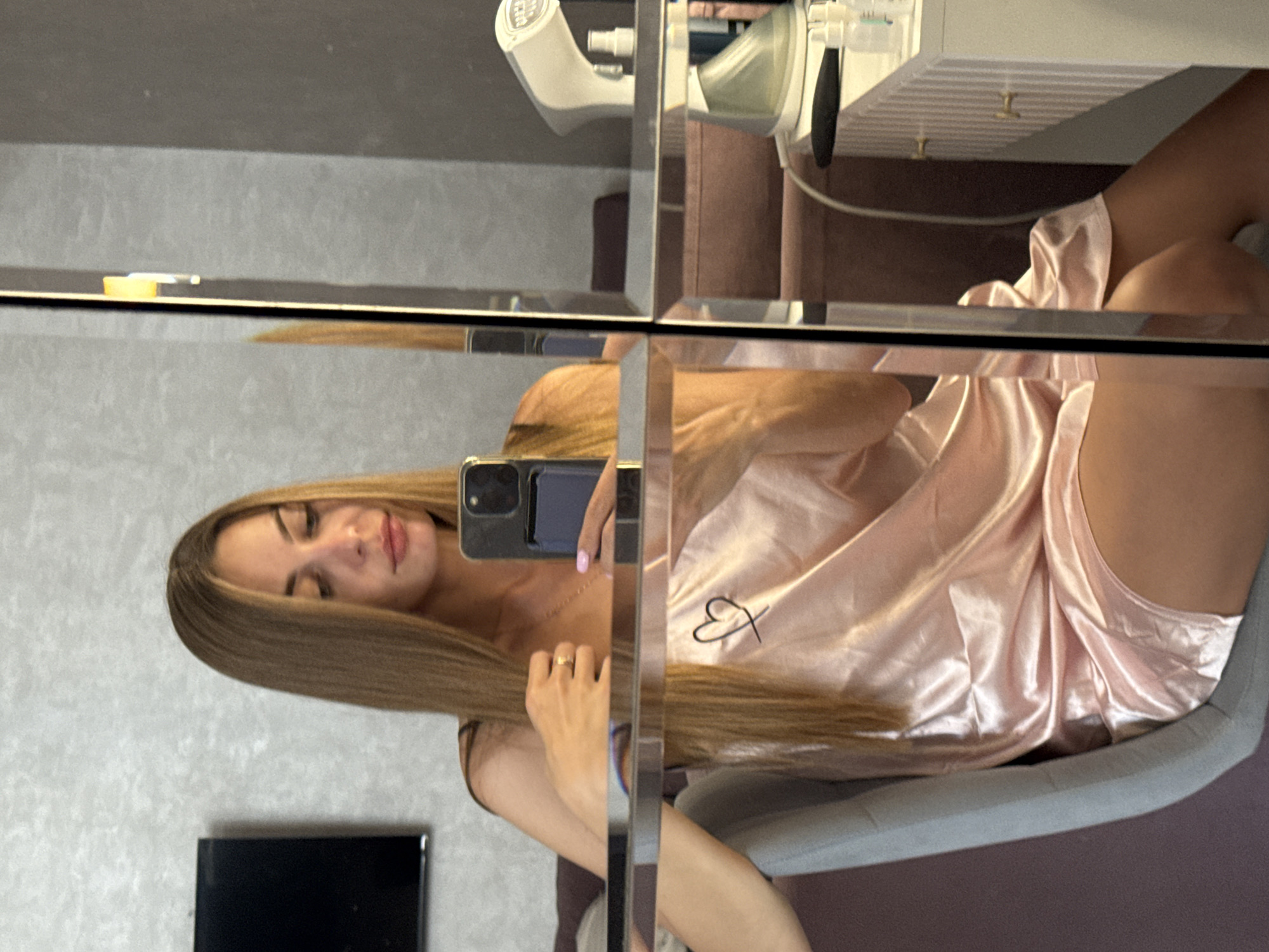 A young woman with long blonde hair is taking a mirror selfie while sitting on a chair. She is wearing a light pink satin slip dress with a small heart embroidered on the left side of the chest. Her right hand is resting on her chest, and her left hand is holding a smartphone. She is wearing a gold ring on her left ring finger and has pink nail polish on her fingernails.
The background shows a modern interior with a textured grey wall and a black television mounted on it. To the right, a mirrored surface reflects a vanity table with what appears to be a garment steamer and some cosmetic bottles. The overall environment suggests a home or hotel room setting. The image does not provide any specific information about the location in Sochi, Russia.