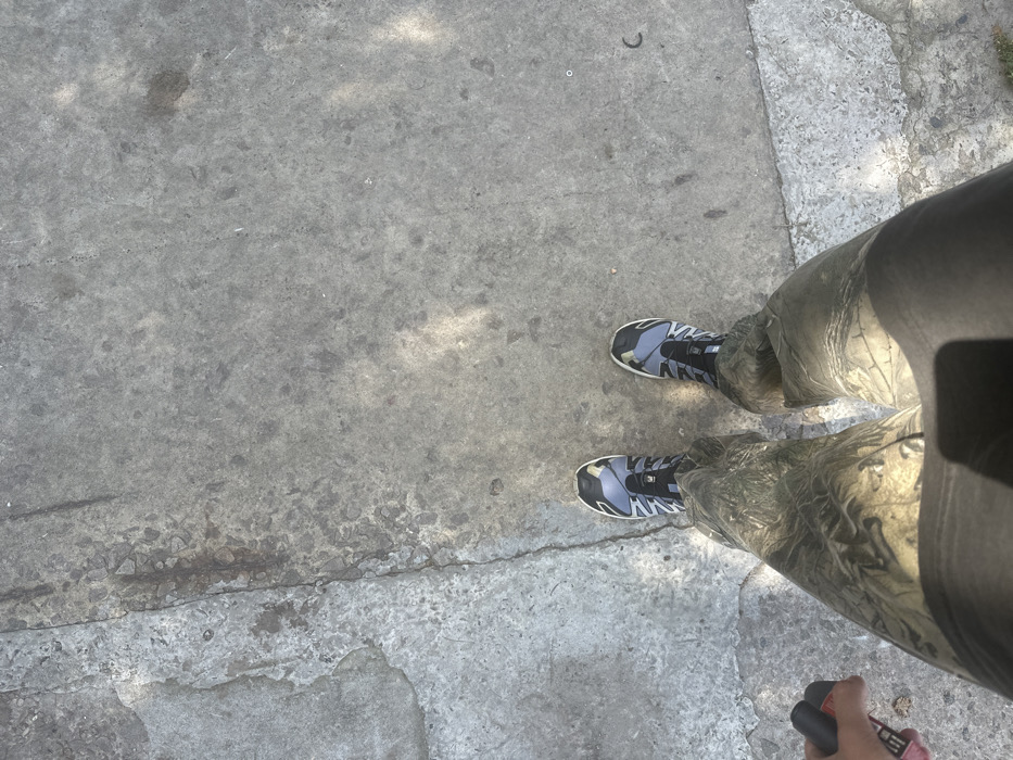 A person is observed from a first-person, downward perspective, standing on a textured, light grey concrete or asphalt surface in Velyka Soltanivka, Ukraine. The ground exhibits irregular cracks, variations in shade, and small embedded aggregates. The individual is wearing camouflage-patterned trousers, primarily in tones of green and brown. Their feet are clad in athletic shoes, predominantly dark grey and light blue, featuring a black quick-lacing system and light-colored midsoles. In the lower-left quadrant of the frame, a hand holds a small, rectangular object with red and black coloration. Sunlight casts irregular patterns of light and shadow across the pavement. To the right of the individual's feet, a small, circular metallic-appearing object and a dark, curved, elongated piece of debris are visible on the ground.