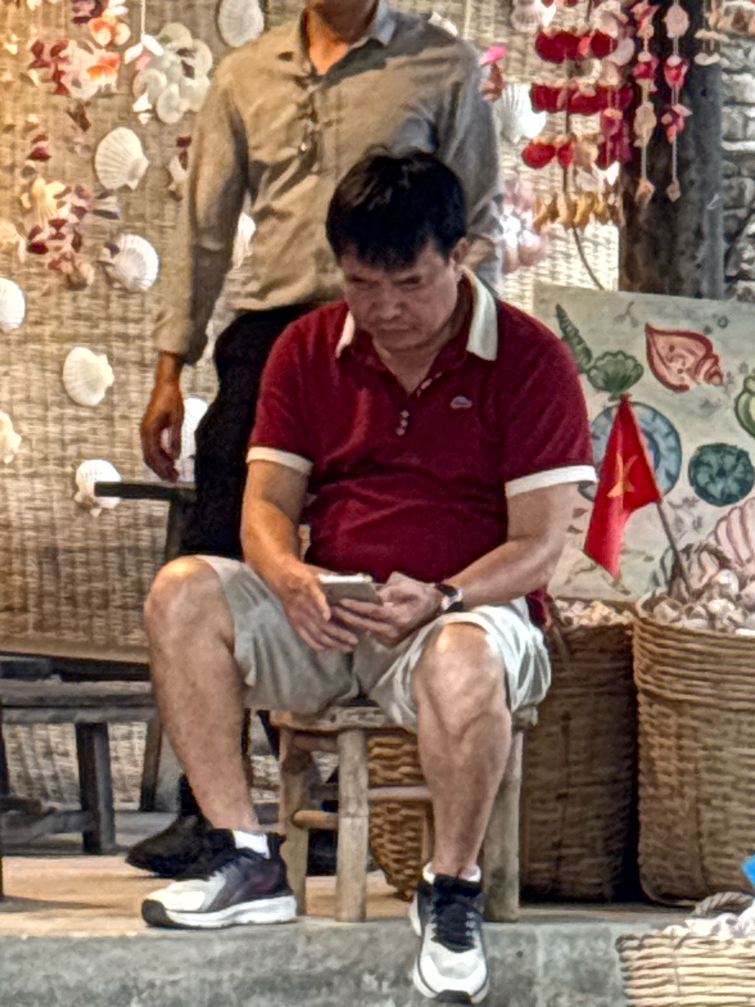 A man wearing a red polo shirt and light-colored shorts sits on a wooden stool, holding a mobile phone. Another person, partially visible, stands behind him, wearing a tan long-sleeved shirt and dark pants. The background features a textured wall decorated with various shells and hanging ornaments, as well as woven baskets. A small Vietnamese flag is planted in a basket to the right of the seated man. The scene appears to be outdoors, possibly in a market or shop, in Nha Trang, Vietnam, given the presence of the Vietnamese flag and the decorative elements.