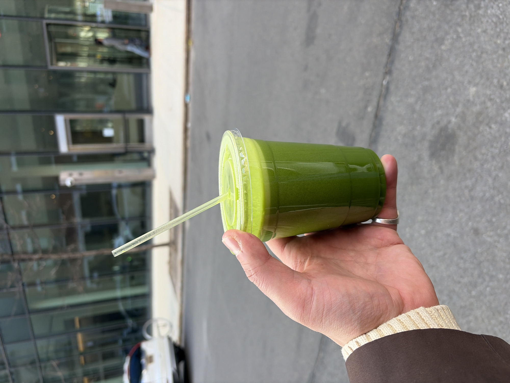 The image shows a hand holding a green juice in a clear plastic cup with a straw. The background includes a city street and buildings. The person is wearing a brown jacket with a white ribbed sweater cuff visible. The location appears to be an urban area, possibly in the city of New York.