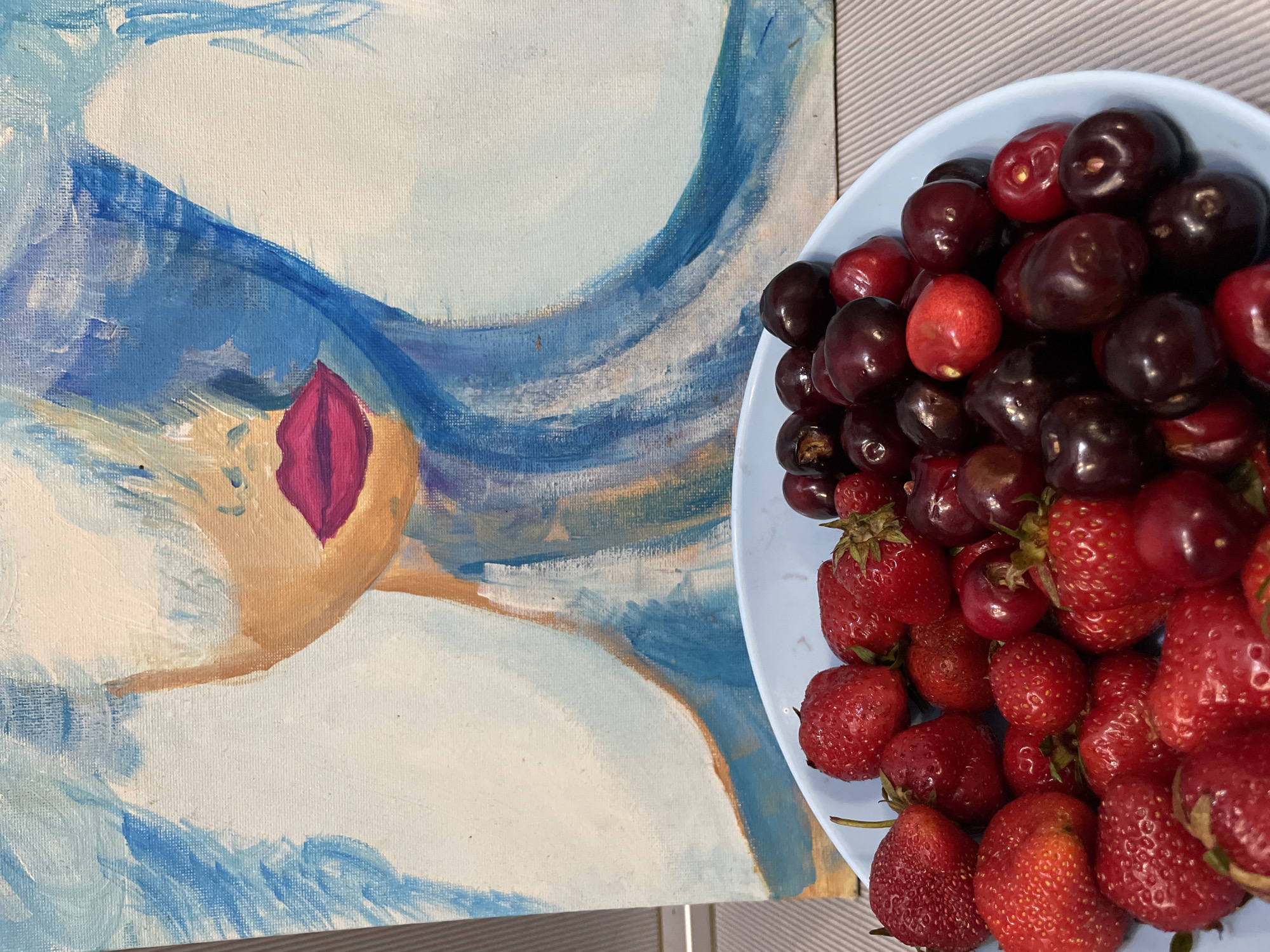 A light blue bowl filled with fresh strawberries and cherries sits on a light gray surface. Behind the bowl, a painting on canvas depicts an abstract portrait of a woman with blue hair and bright pink lips. The painting is positioned on a light gray wall. The overall scene is indoors.