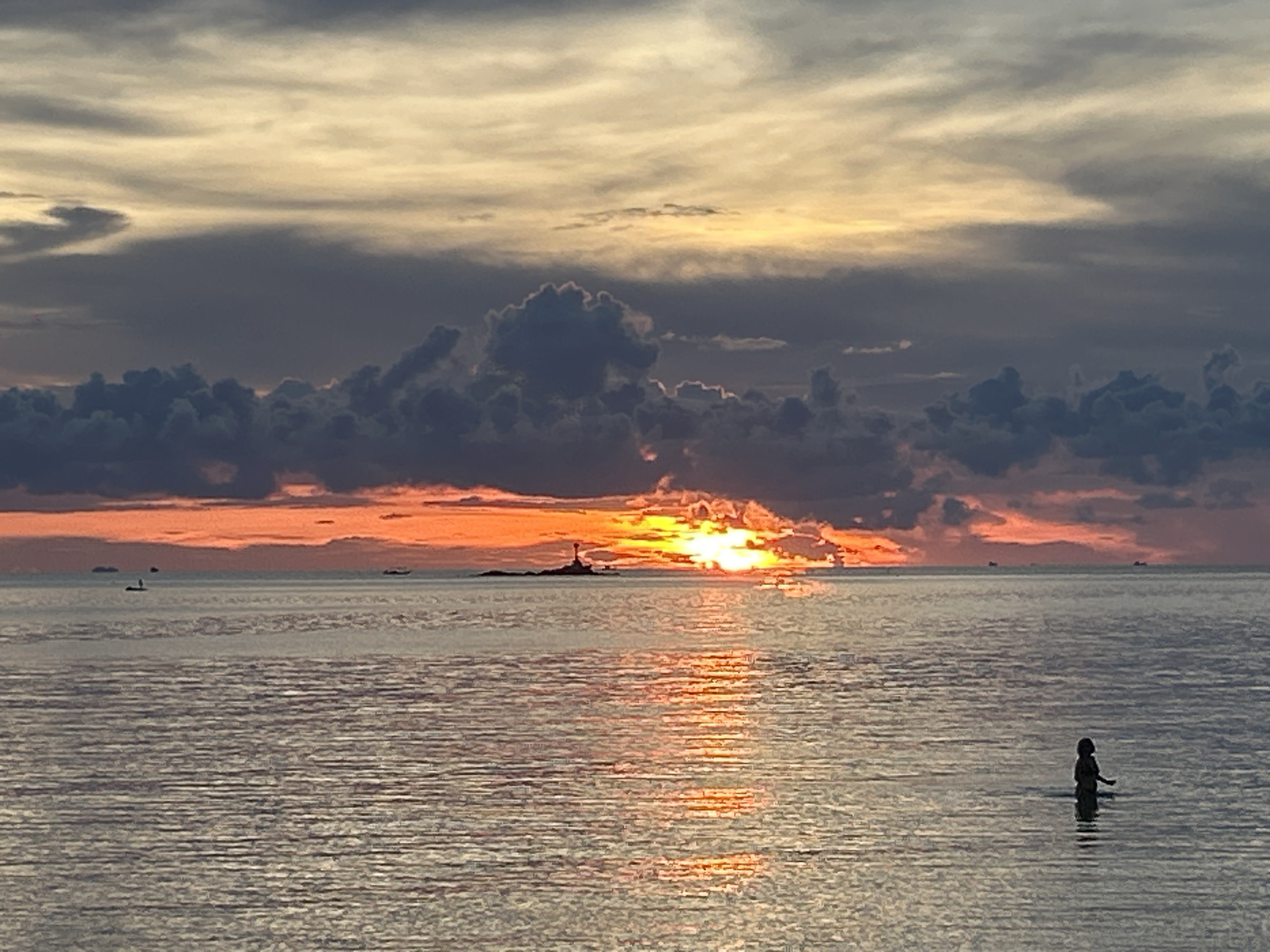 The image captures a sunset over the ocean in Baan Sri Thanu, Thailand. The sun is positioned behind a small, rocky island with a structure that may be a lighthouse or tower. Dark, heavy clouds dominate the top portion of the frame, contrasting with the orange and yellow hues of the setting sun. The sun's light reflects on the surface of the water, creating a shimmering path toward the viewer. In the lower right of the frame, there is a silhouette of a person standing in the water. Several other vessels are visible in the distant background.