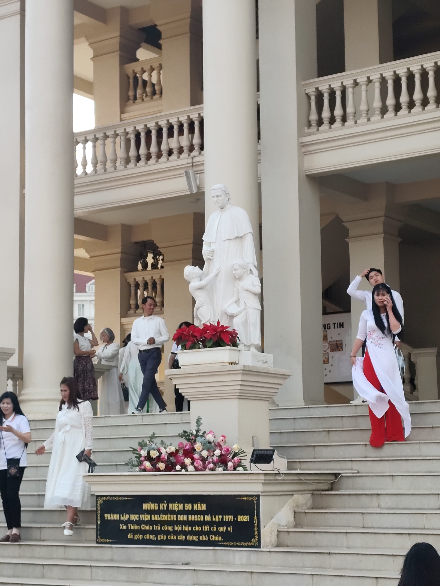 The image shows the exterior of a building, likely a religious institution, with multiple columns and balustrades. A statue of a robed figure with two children stands prominently on a pedestal, adorned with poinsettias. A floral arrangement sits in front of a plaque, which reads "MỪNG KỶ NIỆM 50 NĂM THÀNH LẬP HỌC VIỆN SALĖDIÈNG DON BOSCO ĐÀ LẠT 1971 - 2021 Xin Thiên Chúa trả công bội hậu cho tất cả quý vị đã góp công, góp của xây dựng nhà Chúa.", indicating a celebration of the 50th anniversary of the Salesian Don Bosco Academy in Da Lat, Vietnam.
Several people are present on the steps leading to the building. One woman in a white dress and red pants appears to be on her phone. Another woman, wearing white, is walking down the stairs. Additional people are visible in the background. The environment appears to be outdoors, possibly during daylight hours.